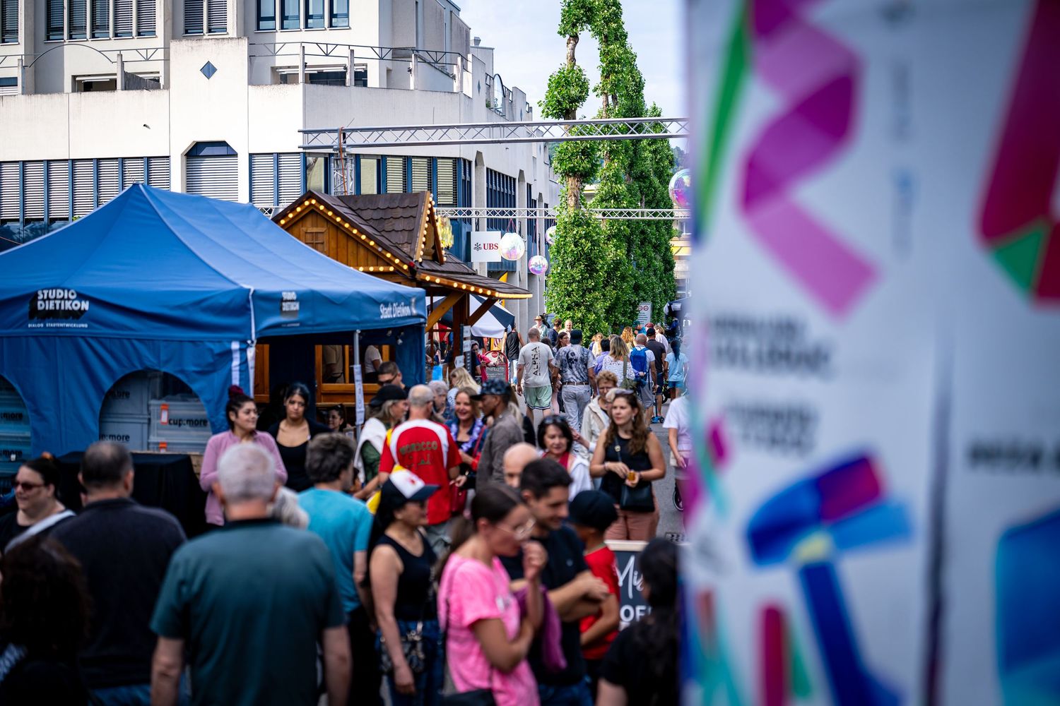 Crowded outdoor street festival with blue tents, vendors, and colorful banners on a busy urban pedestrian walkway.