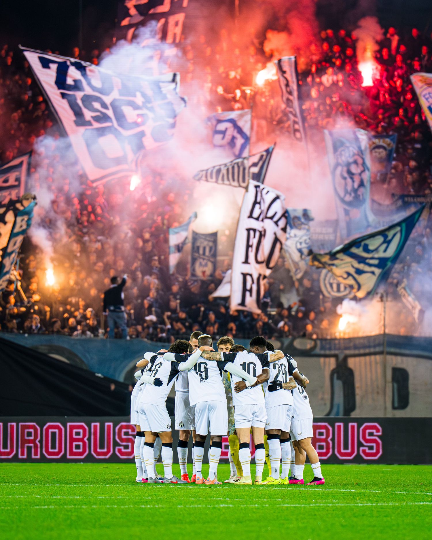 Soccer team huddles on field as fans wave banners and light colorful flares in stadium stands behind them.