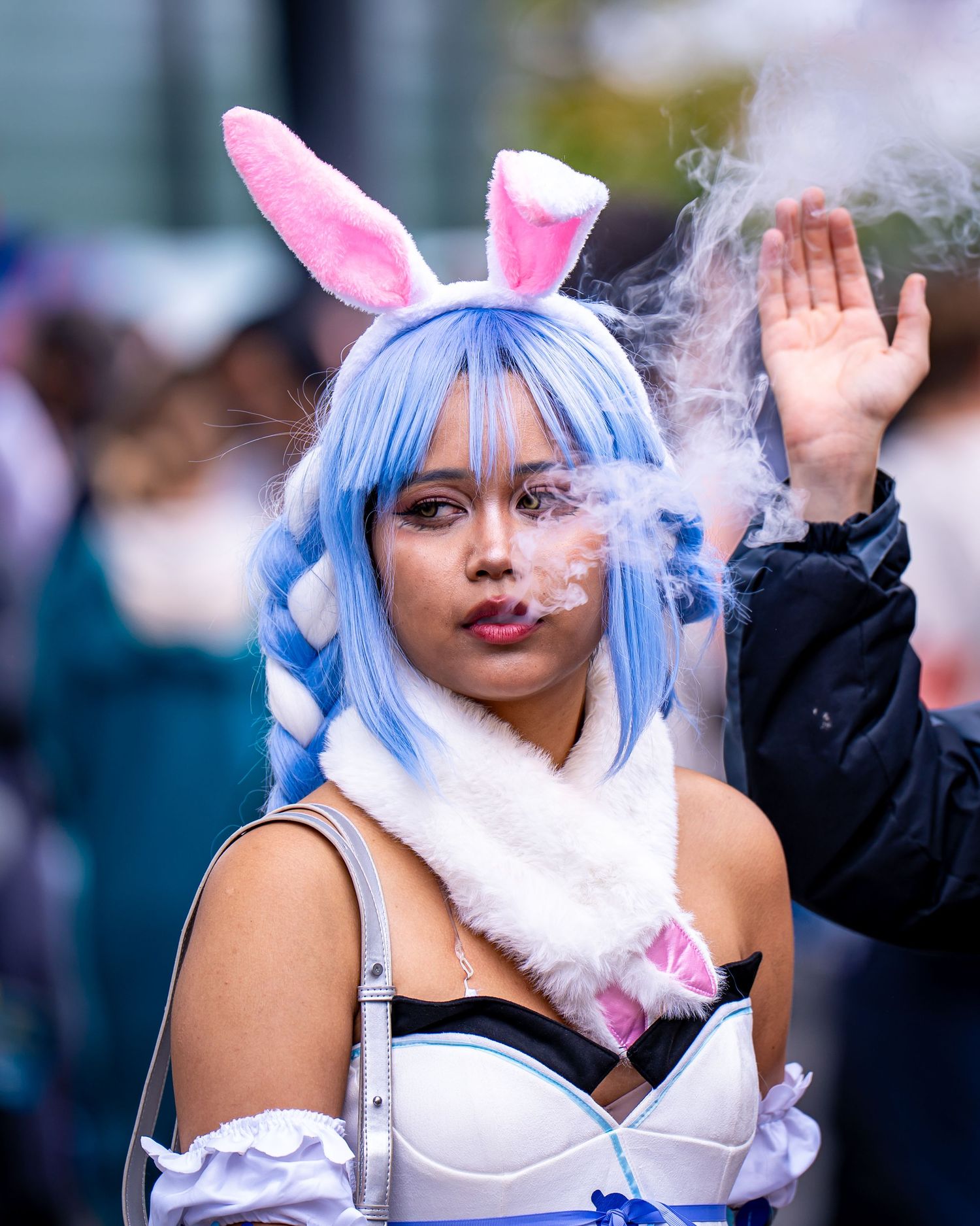 Young woman in blue wig, pink bunny ears, and white cosplay costume exhaling smoke at an outdoor event.