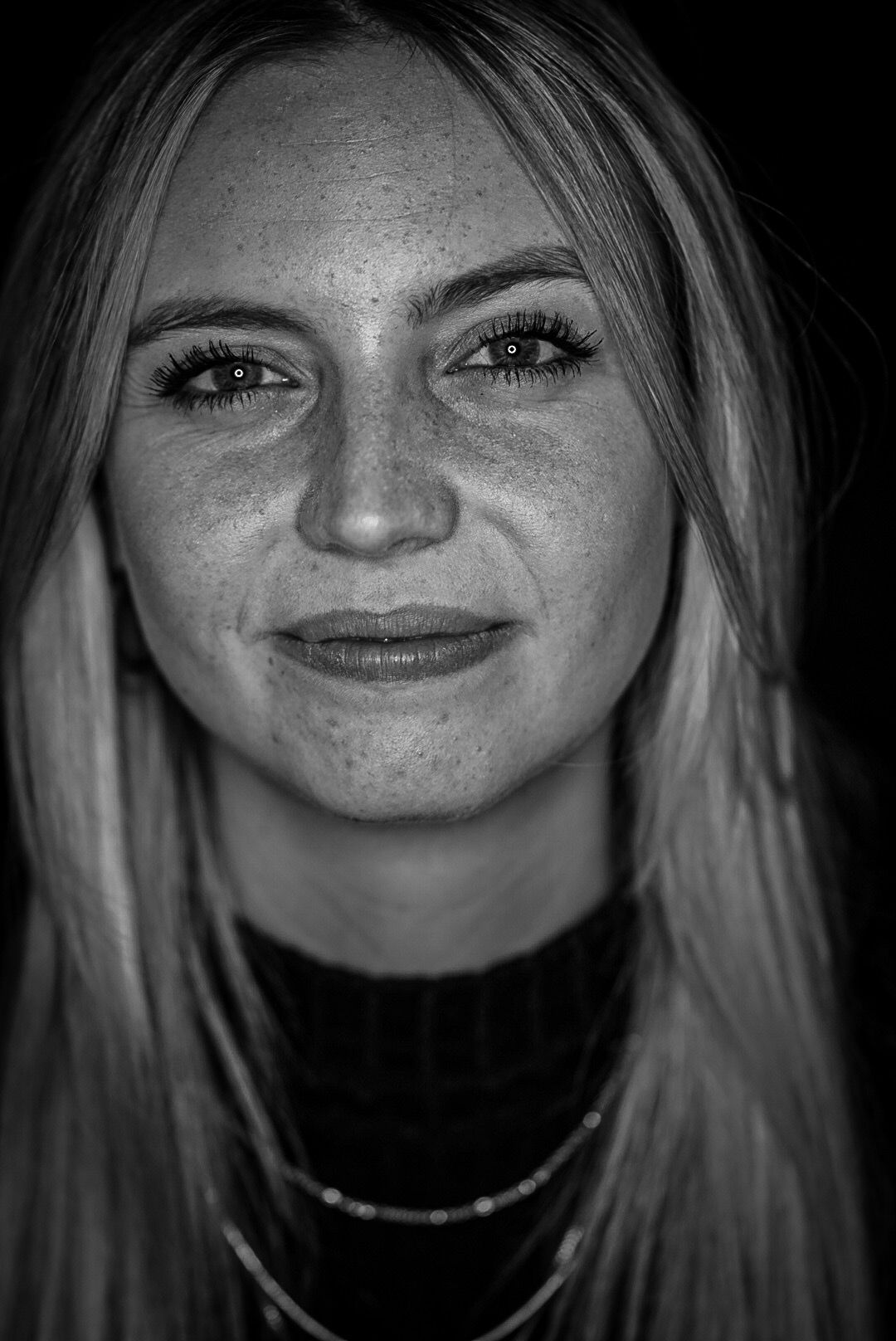 Black and white portrait of a smiling young blonde woman with freckles wearing a dark turtleneck and necklace.