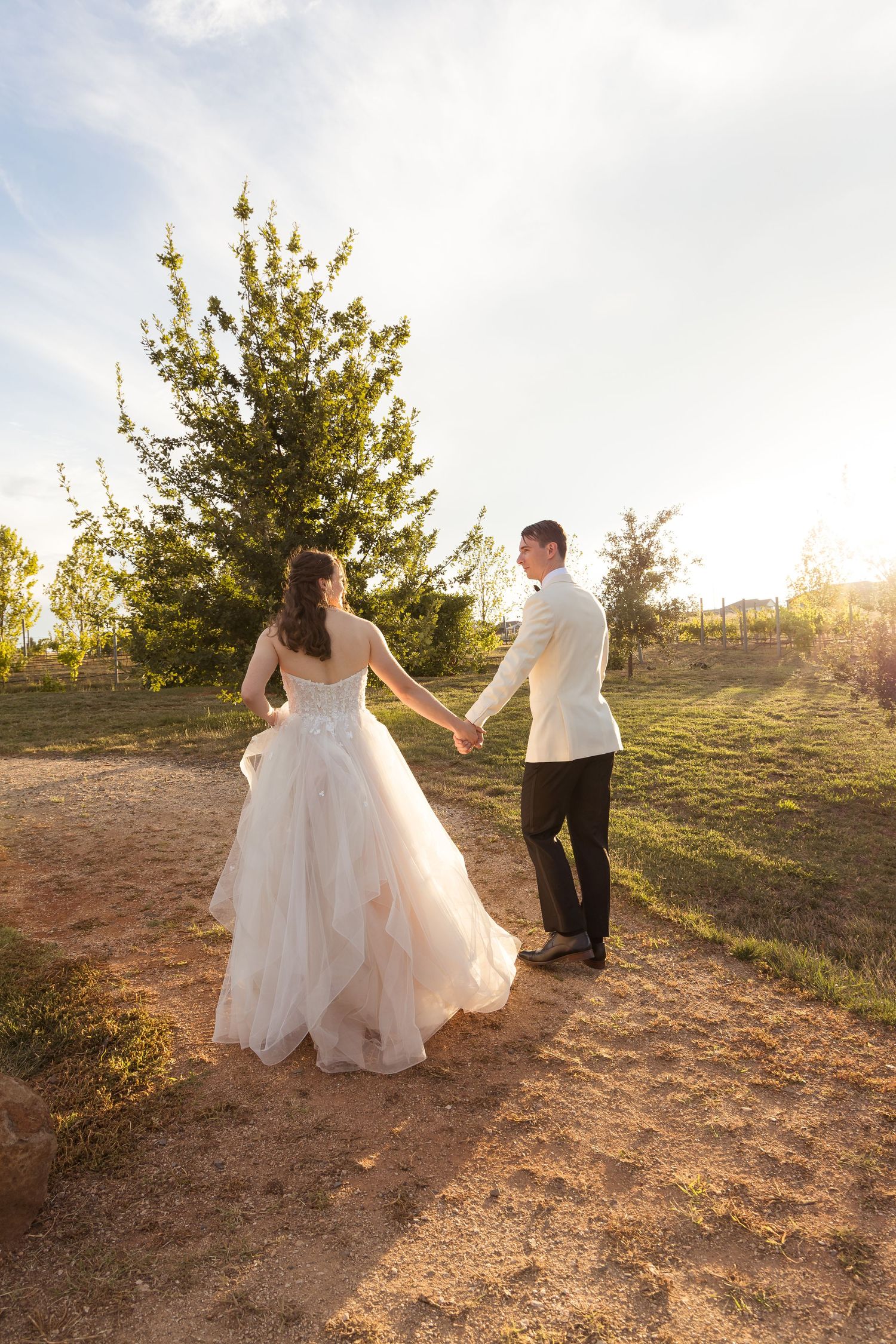 Bride in blush tulle gown and groom in white jacket holding hands walking toward trees in golden sunlight.