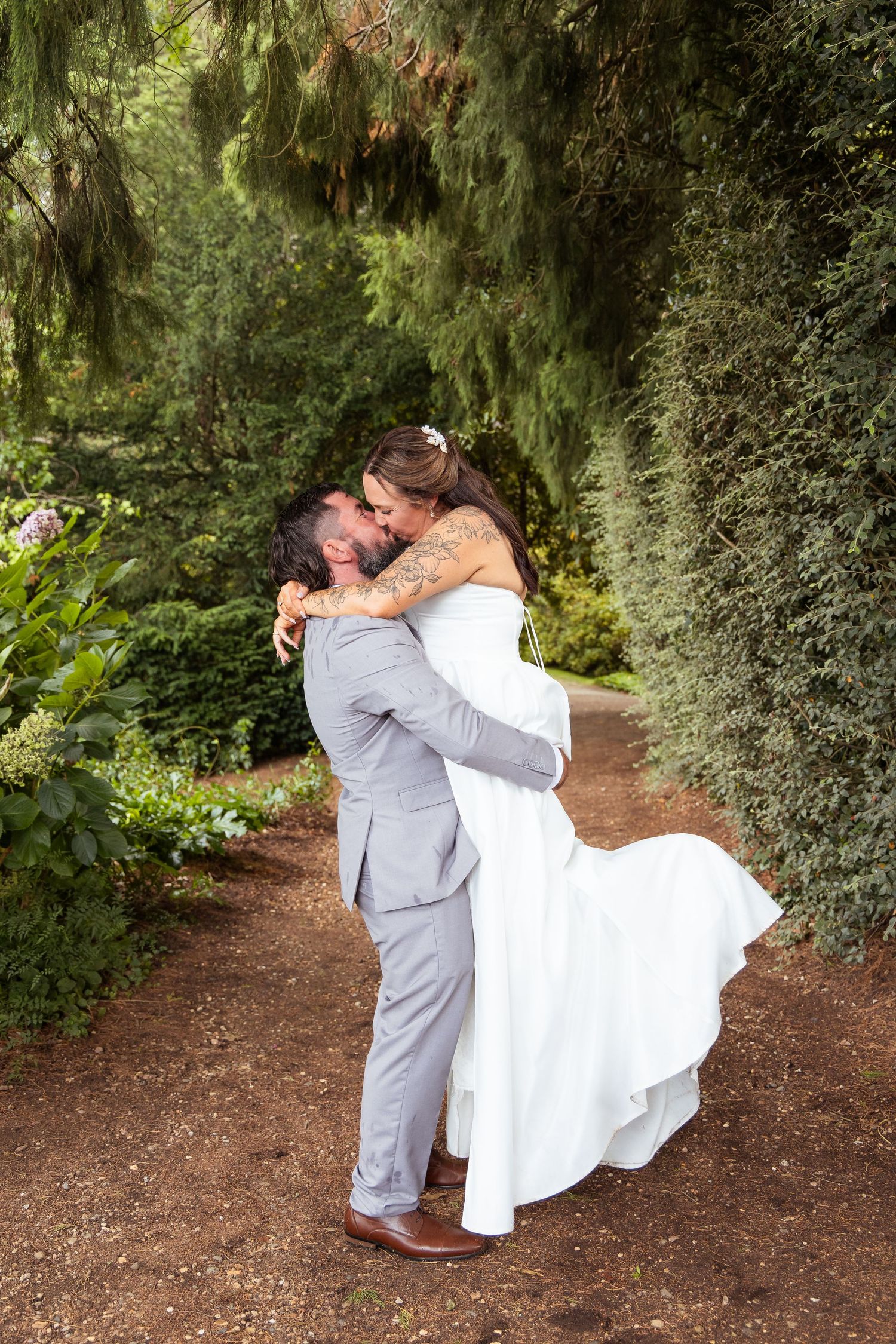 Groom in grey suit lifting bride in white dress while sharing a joyful embrace in lush garden setting.