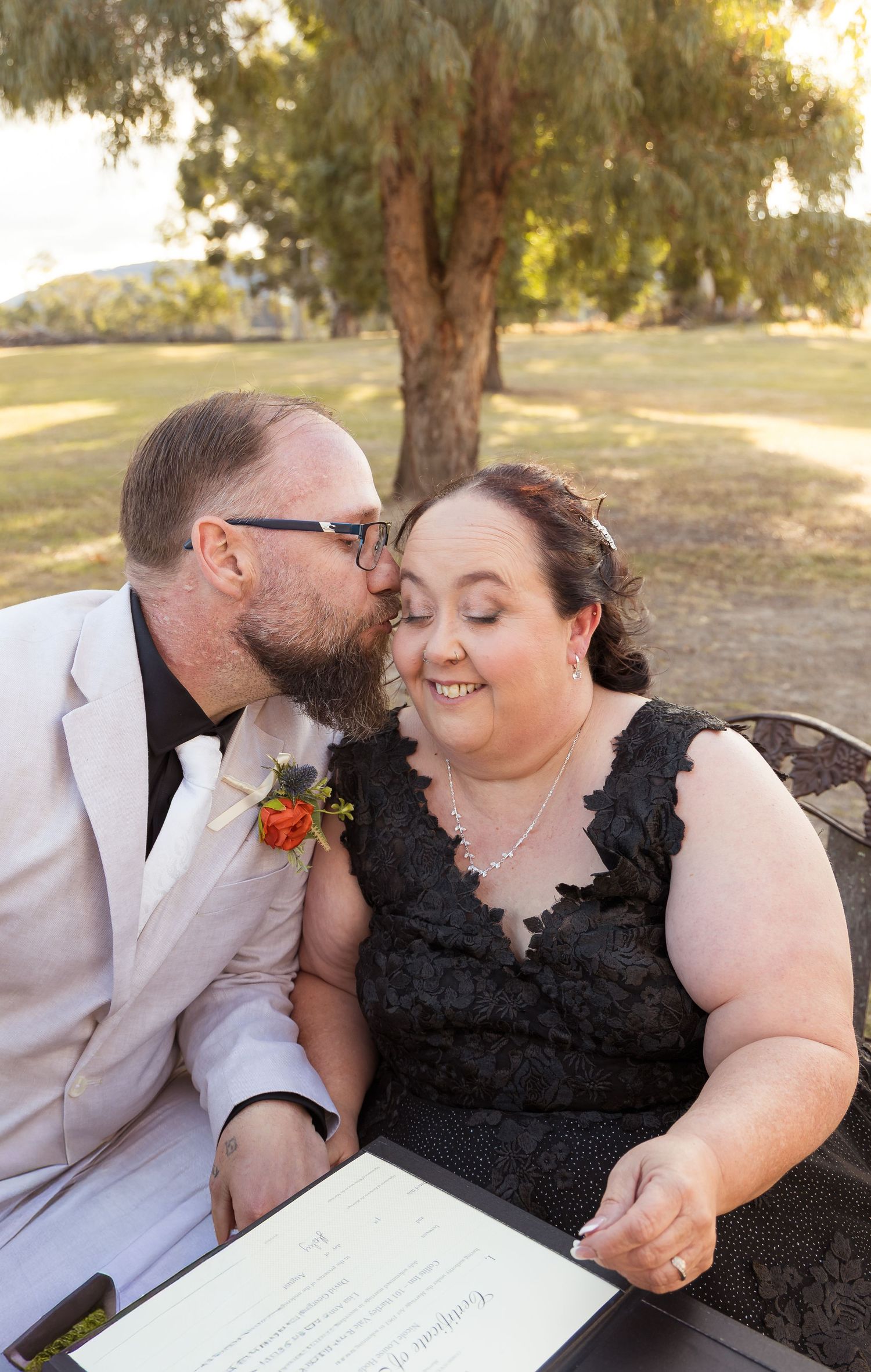 Groom in white suit kisses bride in black lace dress on cheek while signing wedding certificate outdoors.
