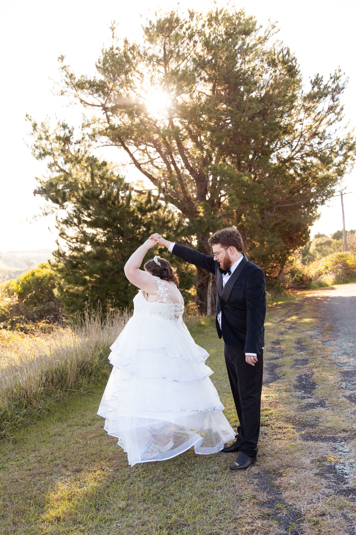 Groom in black tux twirls bride in layered white gown on a sunlit country road beside a large pine tree.