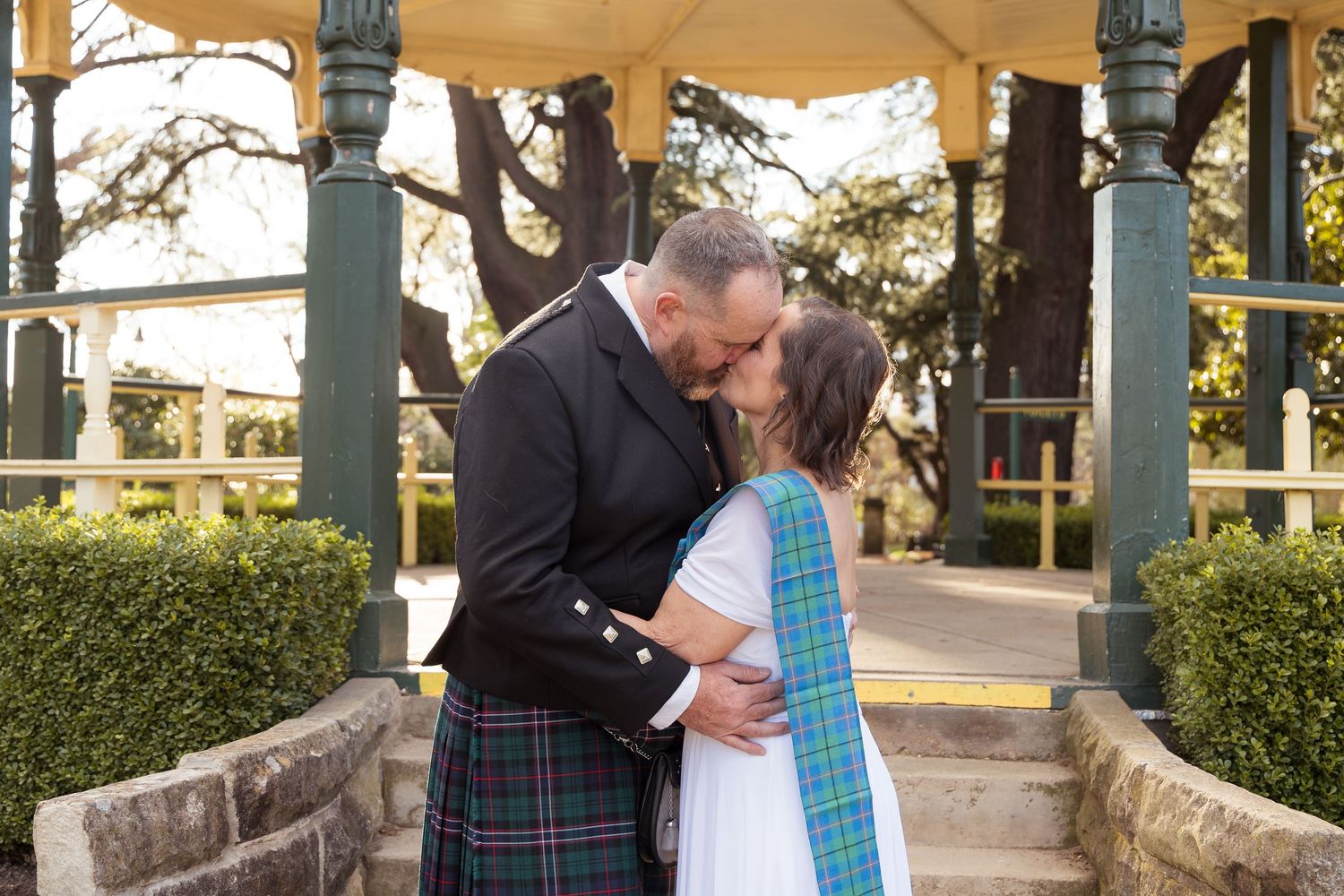 Couple sharing a kiss at gazebo, groom wearing traditional Scottish kilt with tartan sash in garden setting.