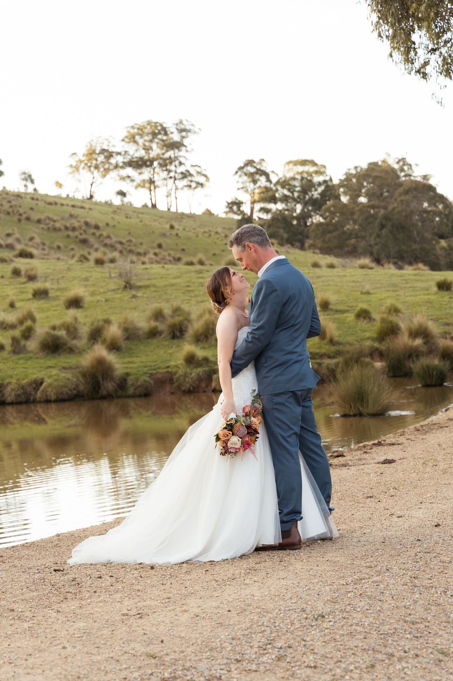 Bride and groom share a romantic embrace by a serene pond in a lush green countryside wedding setting.