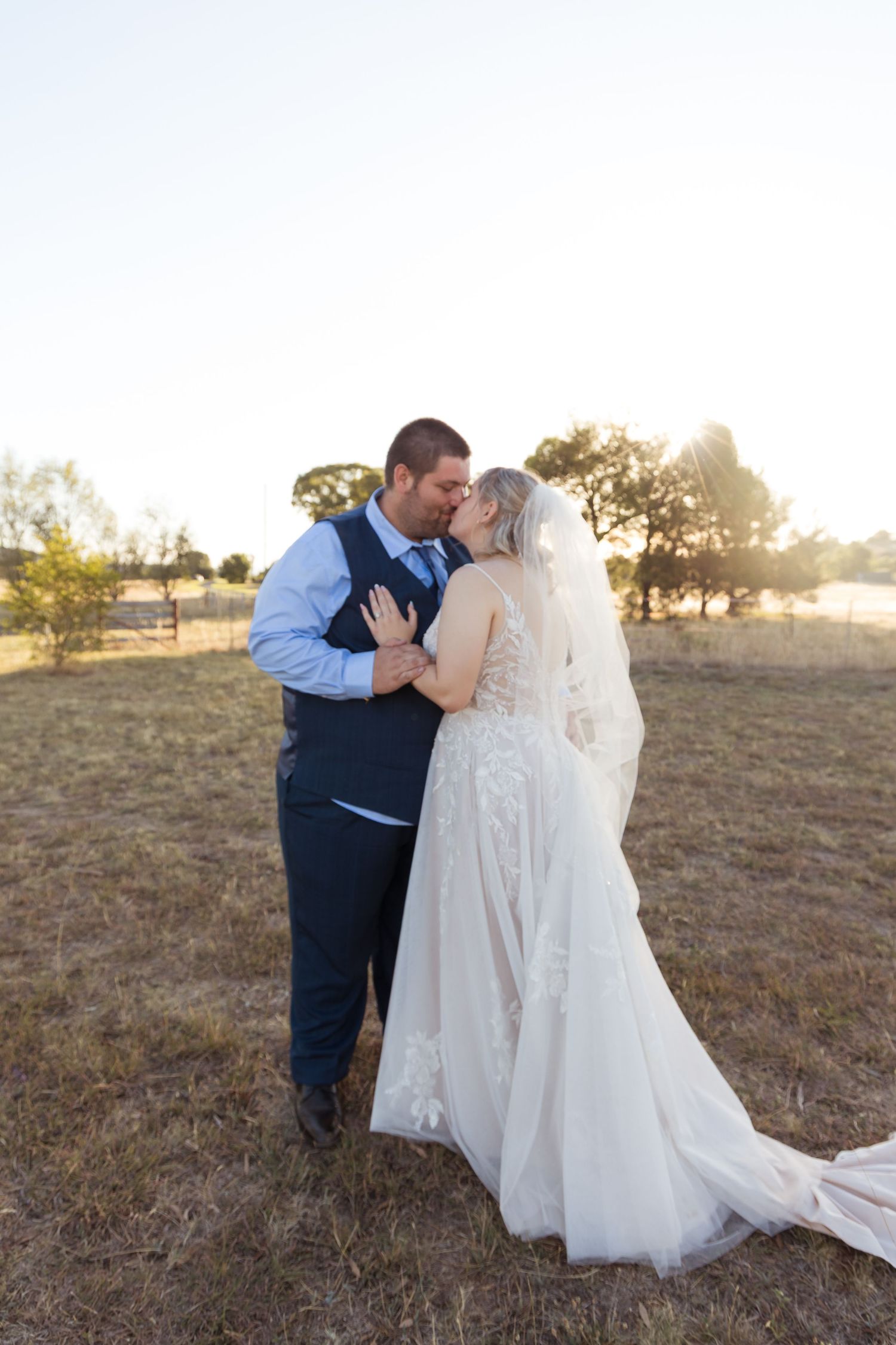 Bride and groom share a romantic kiss at golden hour in an open field, bride wearing a lace gown with veil.