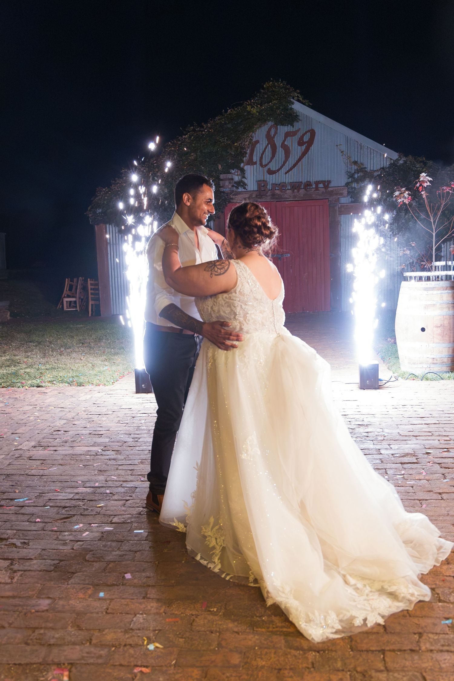 Bride and groom share first dance outdoors at night with sparkling fountains and rustic 1859 barn backdrop.