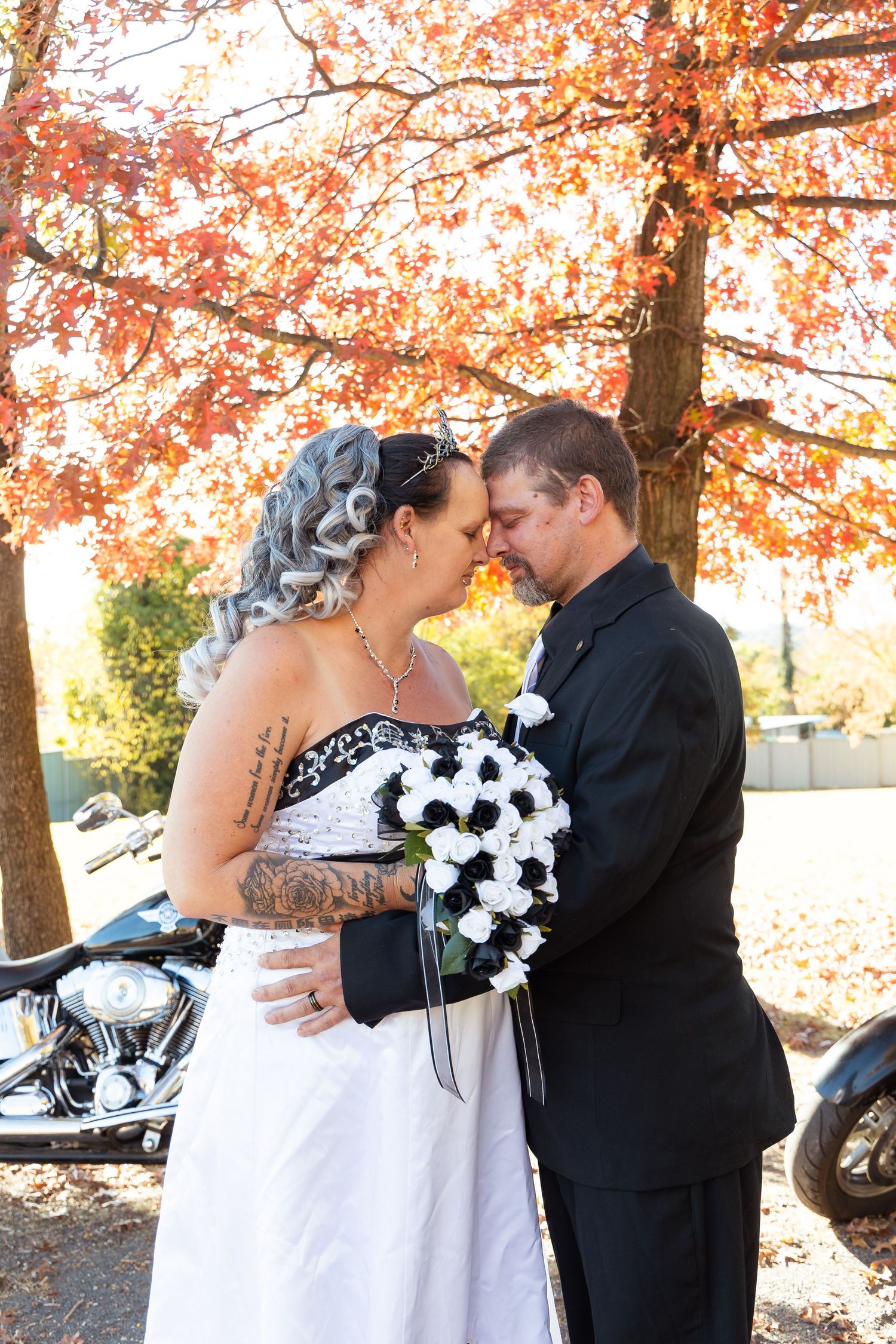 Bride and groom touching foreheads tenderly outdoors, fall foliage backdrop, black and white wedding attire, bouquet of roses.