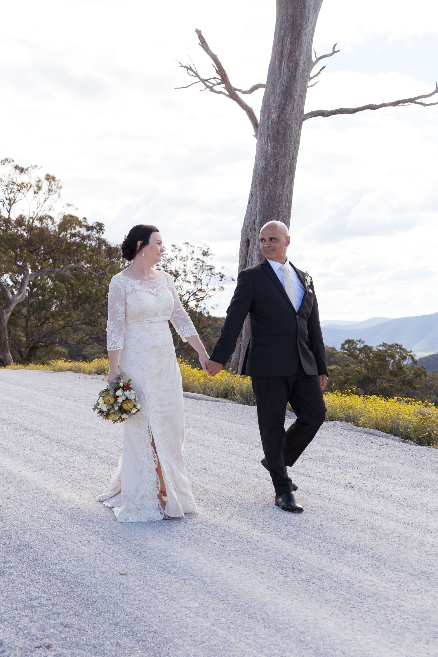 Elegant bride and groom walking hand in hand on rural road with wildflowers and bare tree in background.