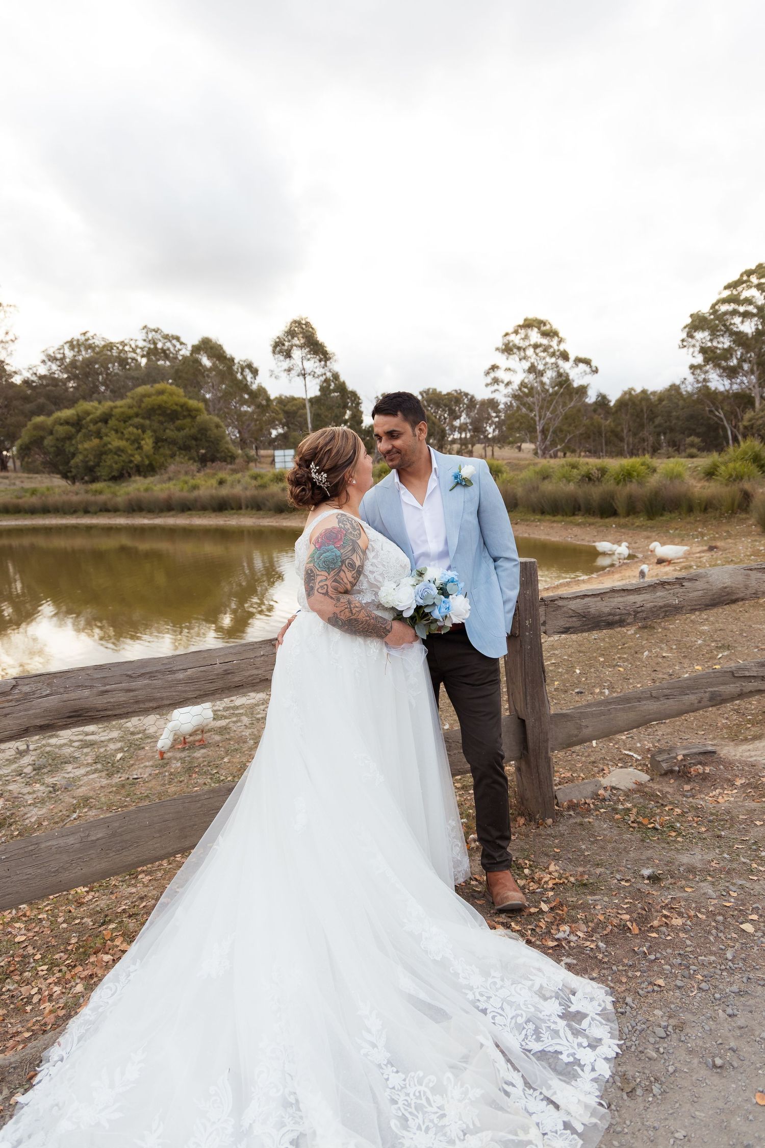 Bride with floral tattoos and groom in light blue suit stand by a tranquil pond holding a blue bouquet.