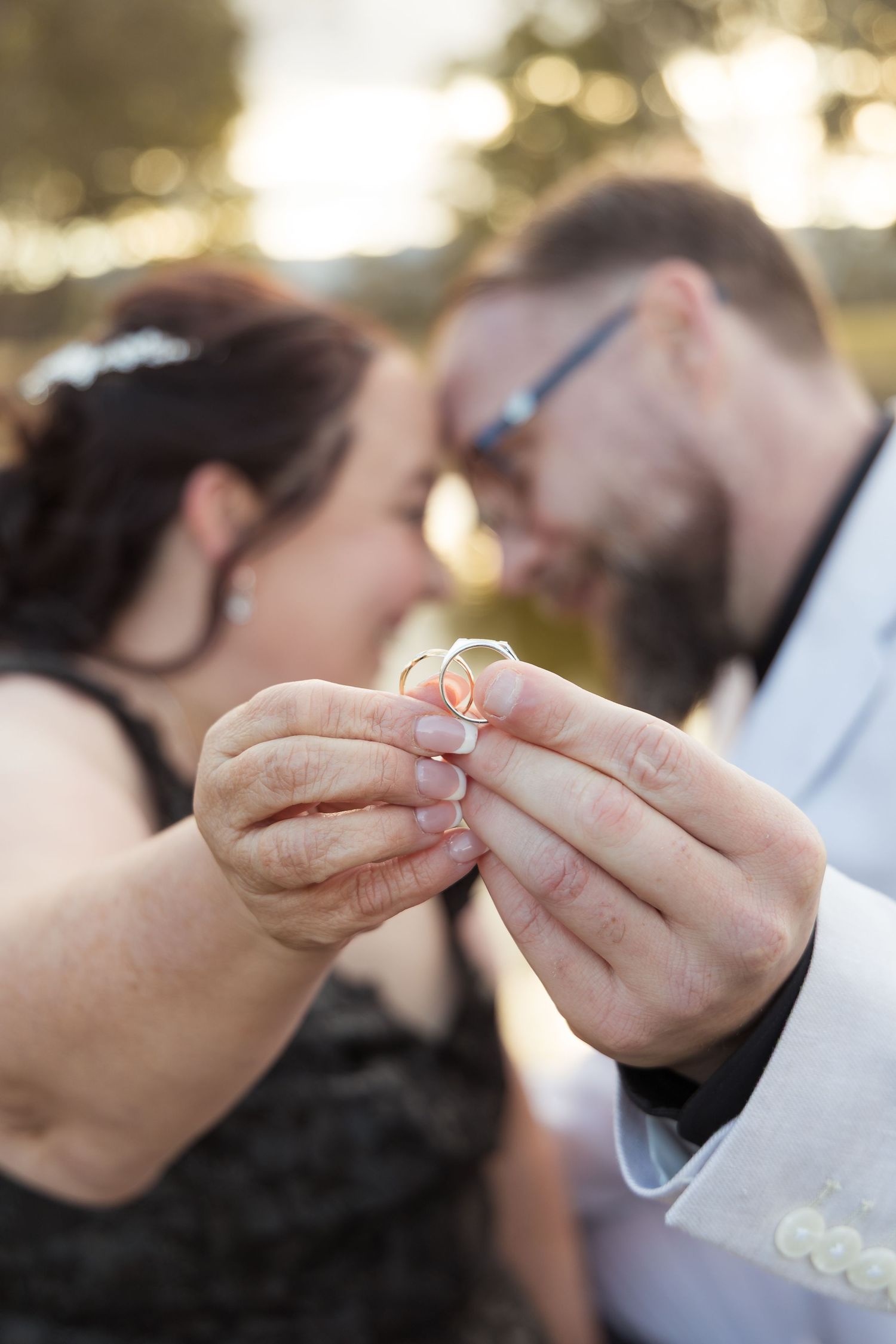 Couple holding wedding rings close together, faces nearly touching, celebrating their engagement outdoors at golden hour.
