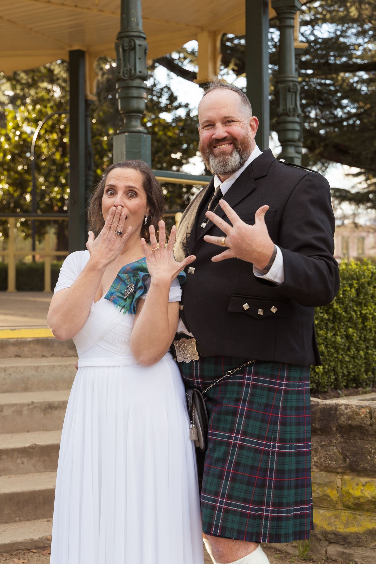 Newlyweds showing wedding rings, woman in white dress, man in Scottish tartan kilt, celebrating outdoors near ornate gazebo.