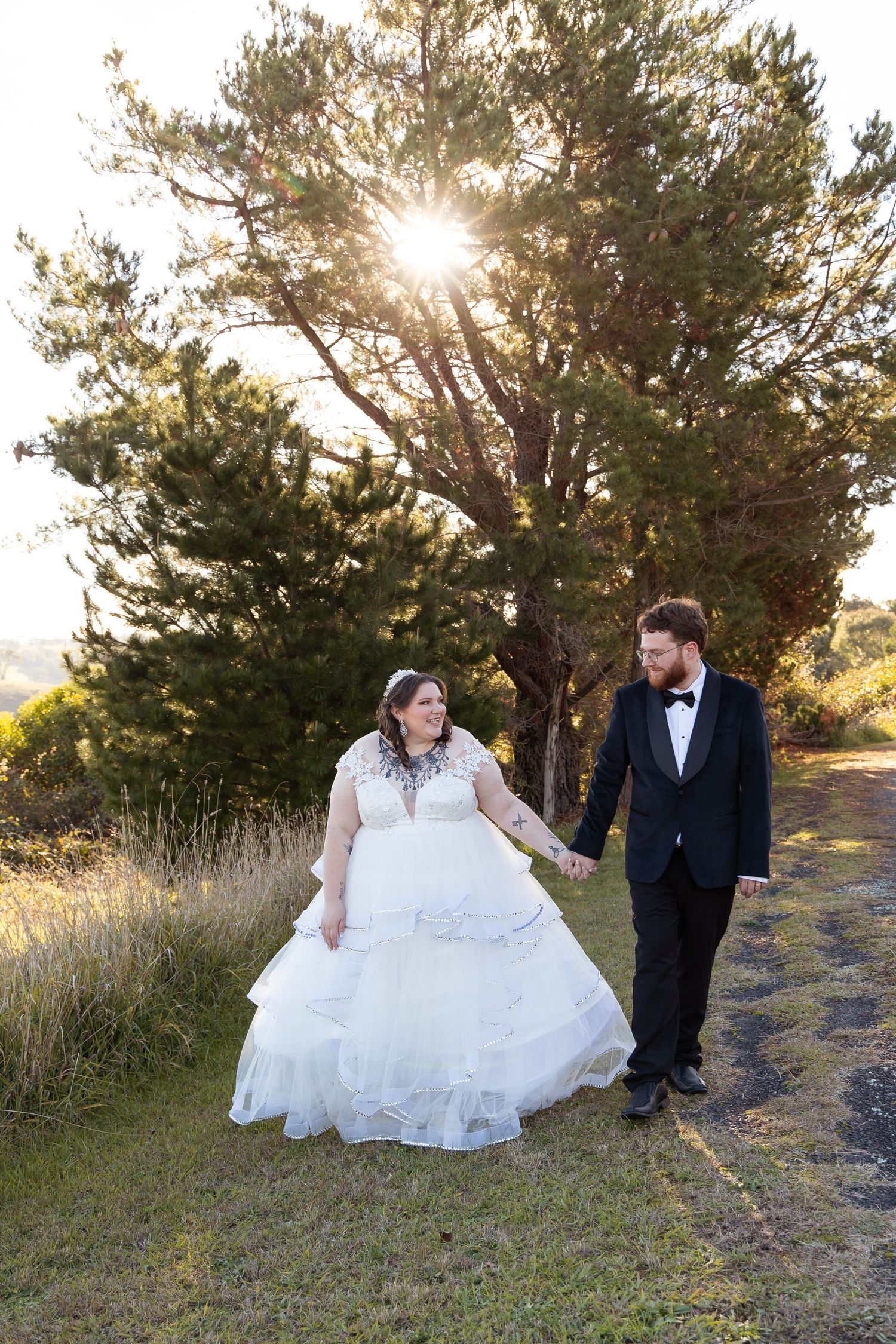 Newlyweds walk hand in hand along a sunlit path with a large tree glowing behind them.