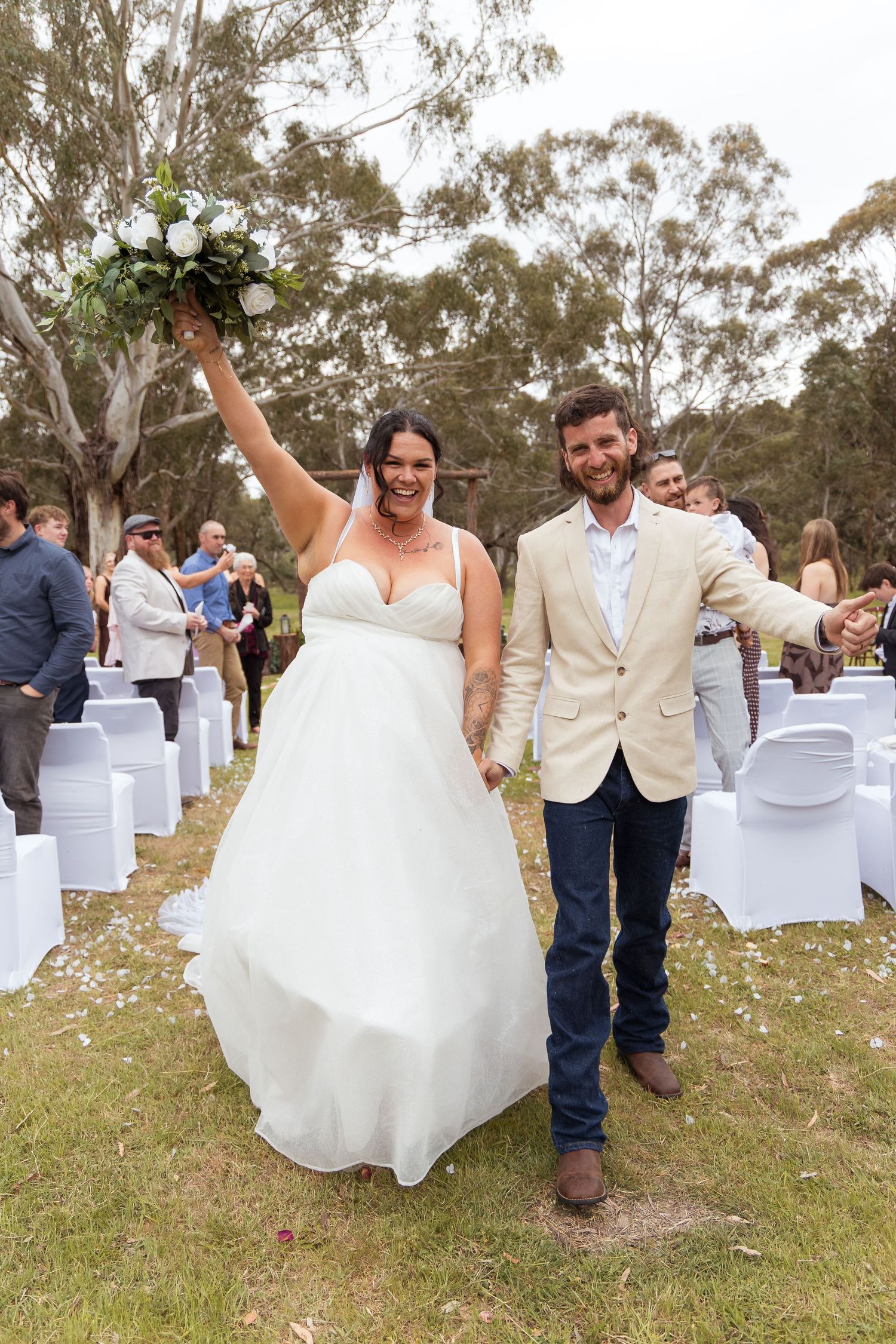 Joyful bride in a white gown raising a bouquet and groom in a cream blazer walk down an outdoor aisle after their bush wedding ceremony.