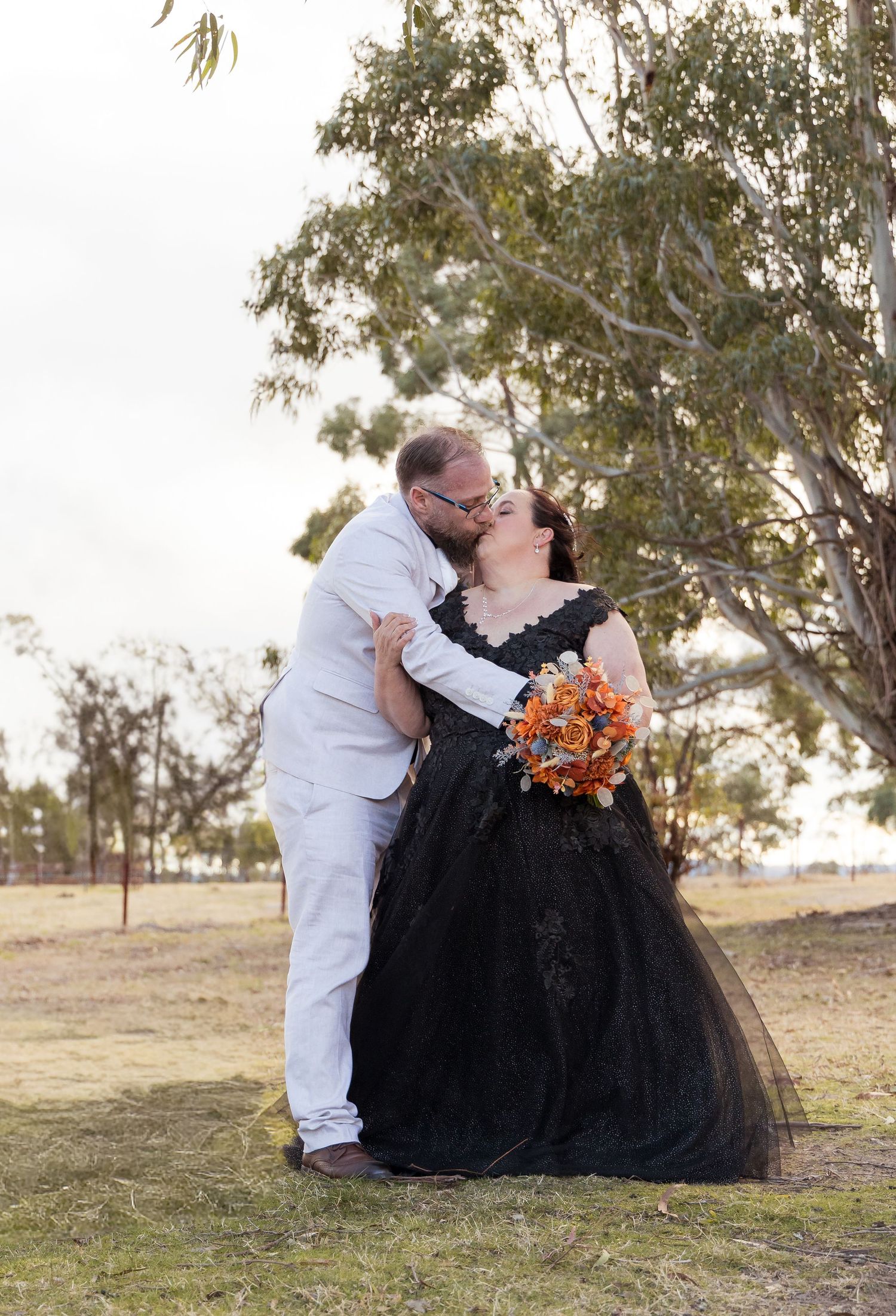 Couple sharing a kiss outdoors, bride in black lace gown holding orange bouquet, groom in white suit, tree backdrop.