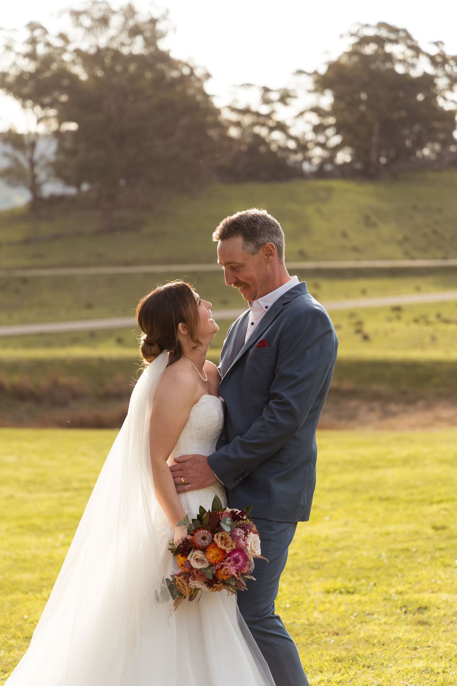 Bride and groom share a romantic moment outdoors at golden hour, bride holding a colorful bouquet in a white gown.