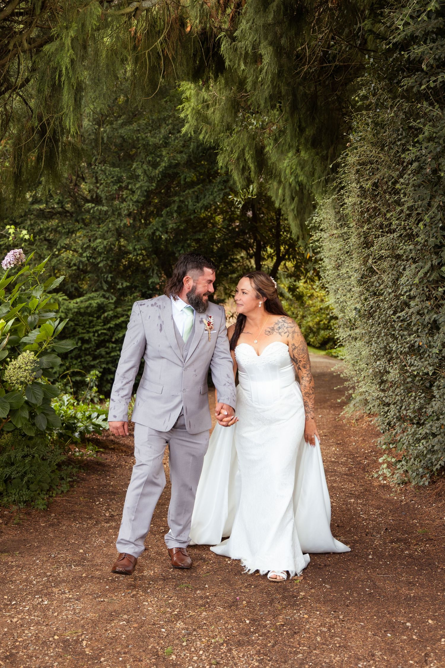 Bride and groom walk hand in hand along a lush garden path, smiling at each other in natural light.