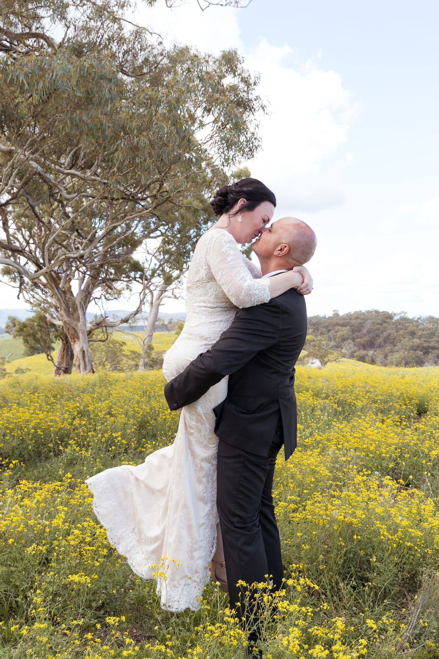 Groom lifts bride in a field of yellow wildflowers for a romantic outdoor wedding portrait.