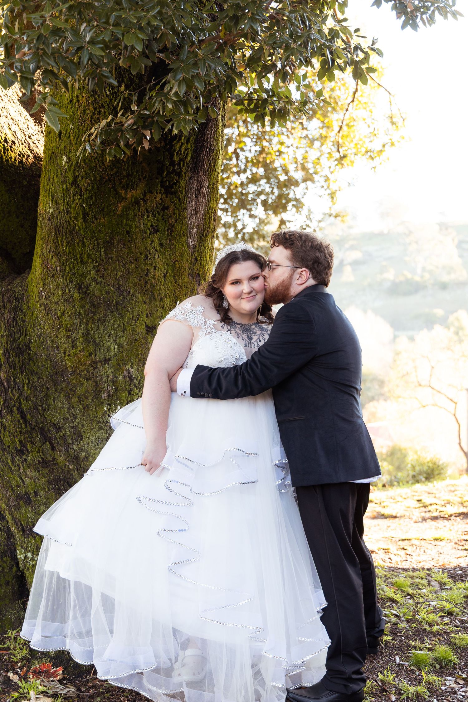 Groom kisses bride on the cheek beside a large mossy tree in a romantic outdoor wedding portrait.