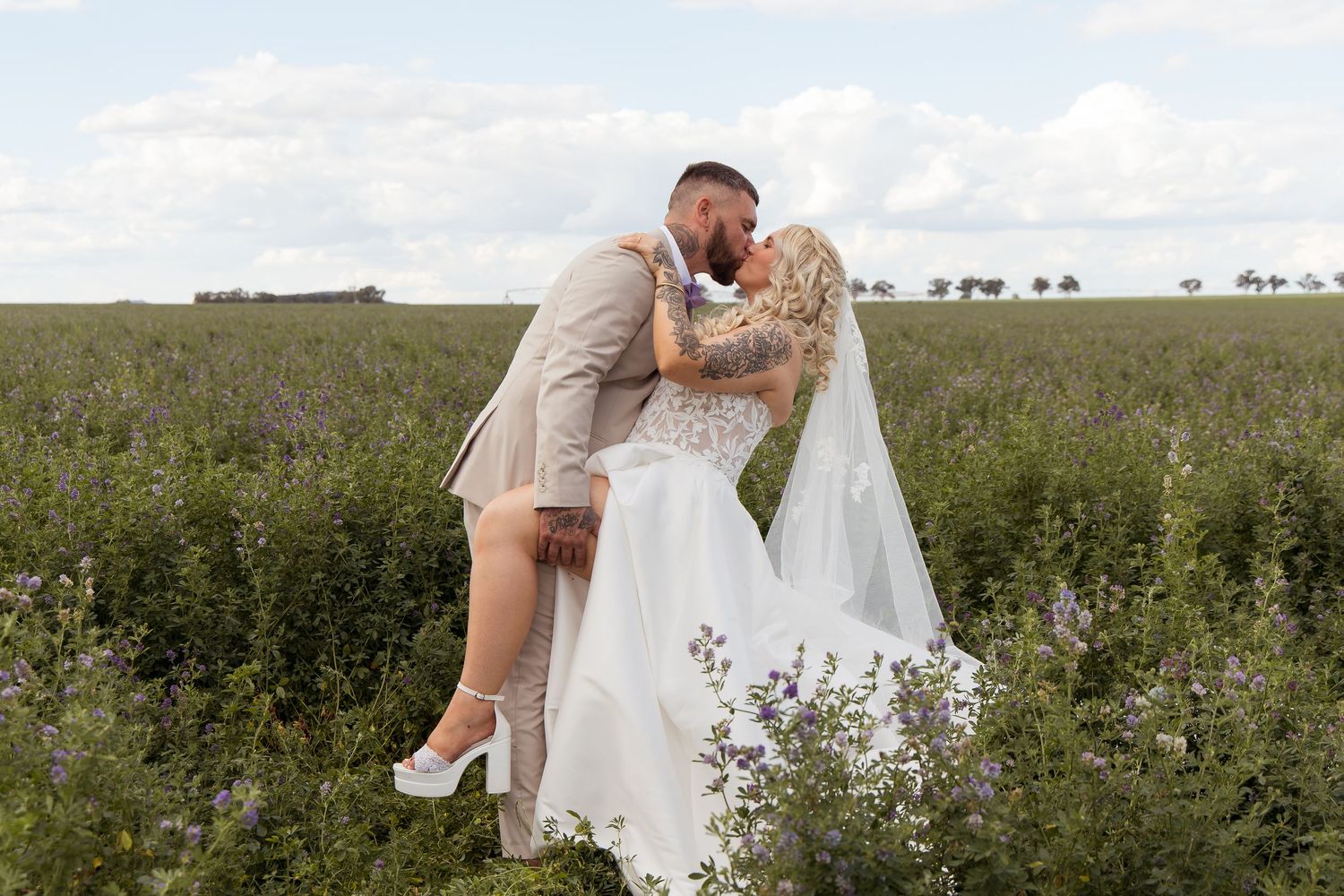 Bride in a white lace gown and veil kisses her groom in a beige suit while dipping back in a green wildflower field under a blue sky.