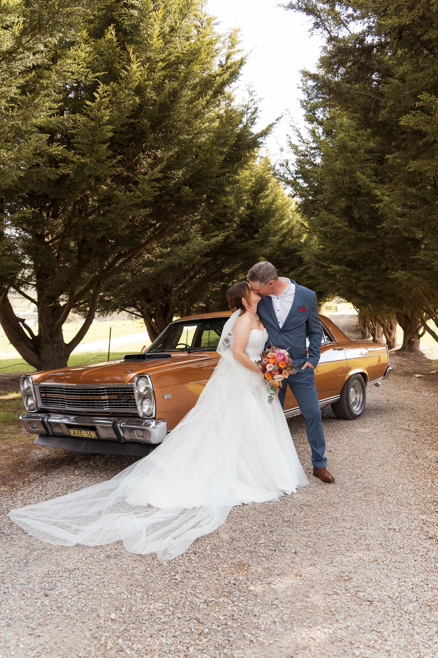 Bride and groom kissing beside a vintage brown car on a gravel driveway lined with tall green trees.