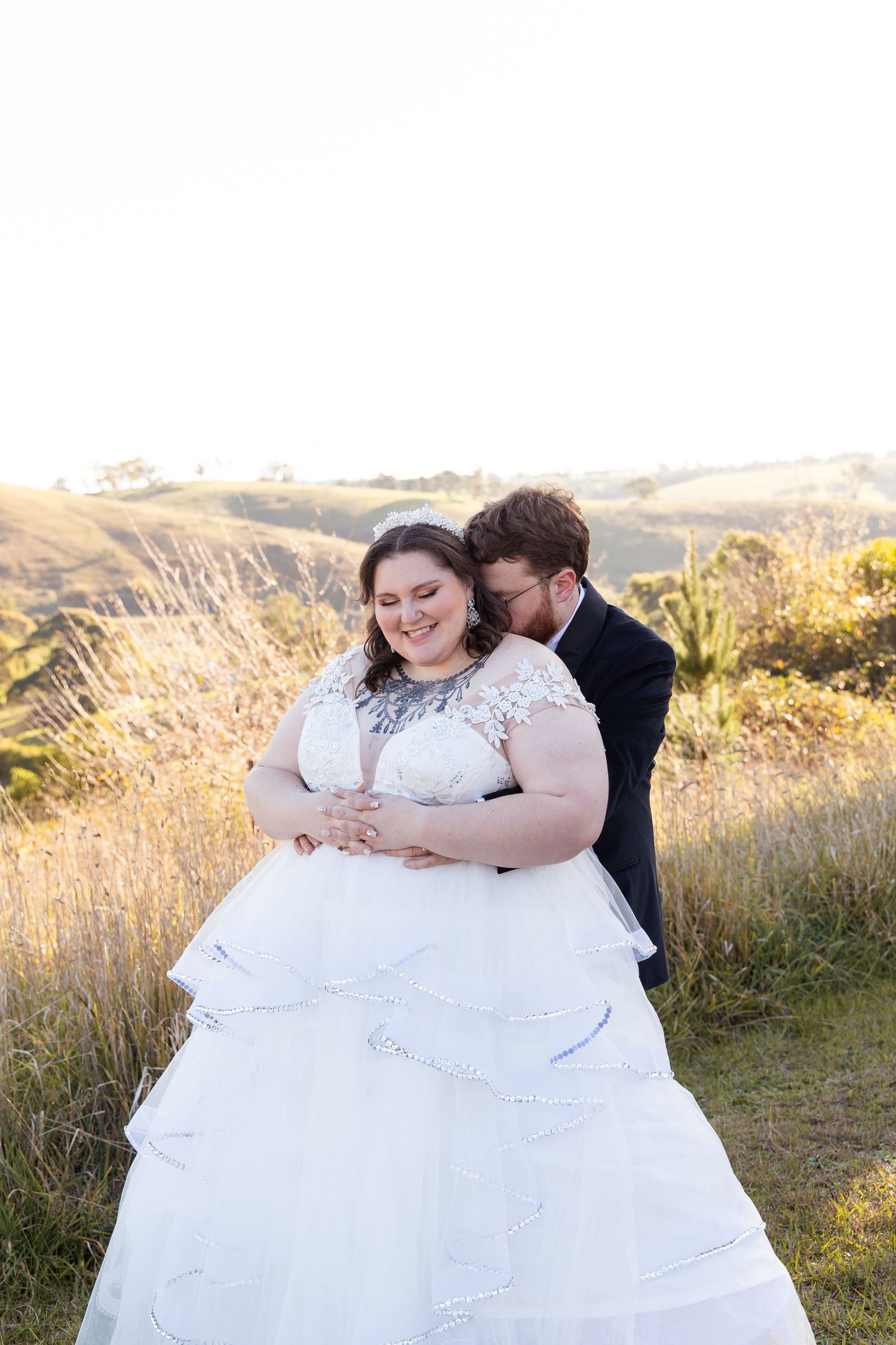 Bride in white tiered lace gown embraced by groom in dark suit, posing in golden sunlit grassy hills.