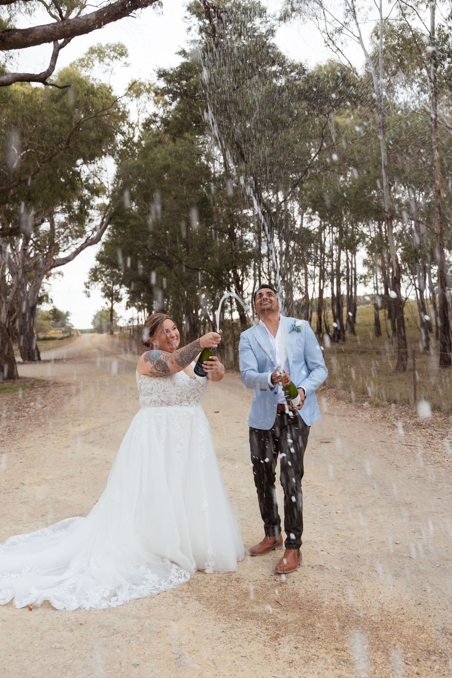 Joyful newlyweds spray champagne on a dusty tree-lined road celebrating their wedding day.