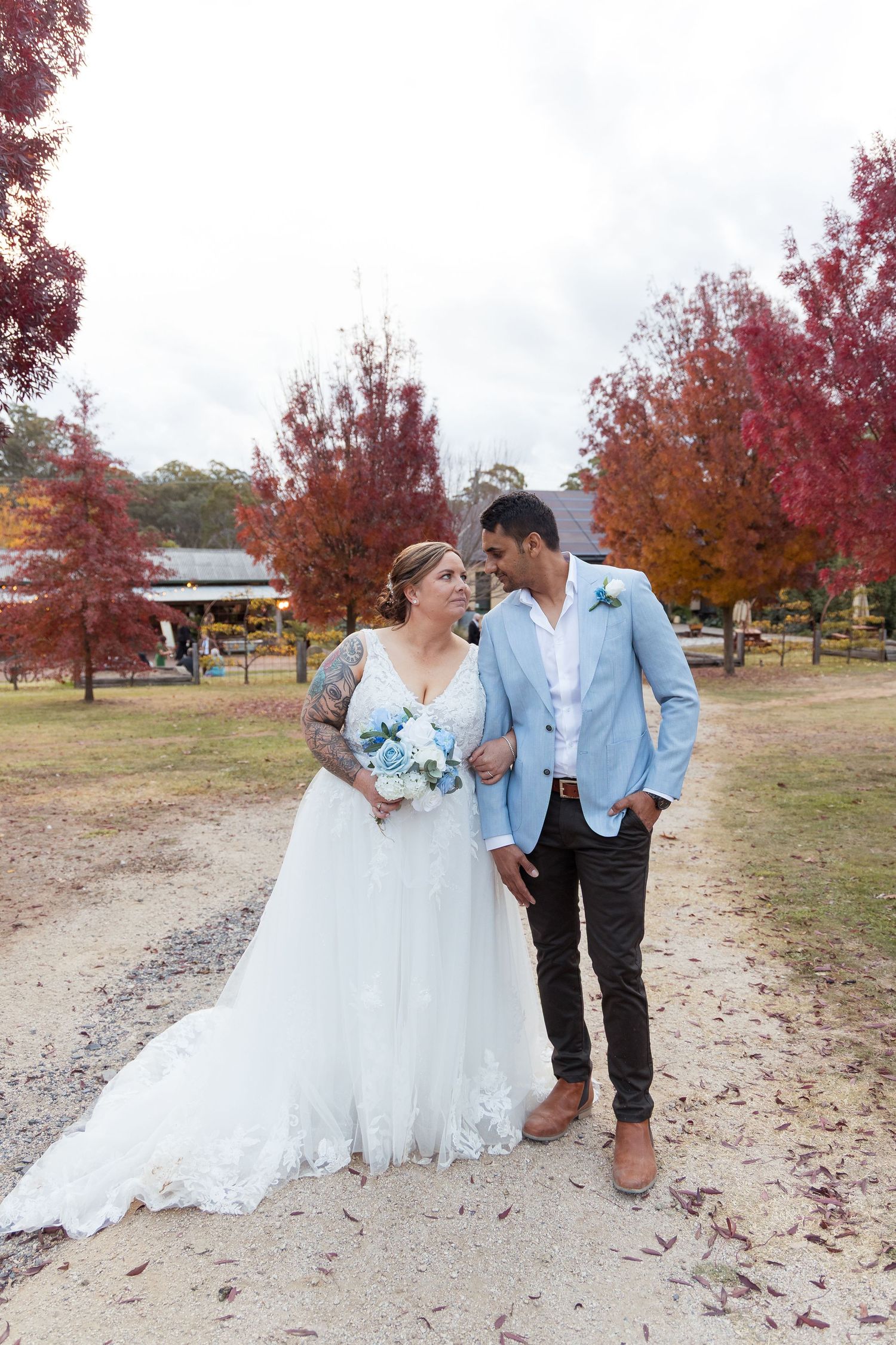 Wedding couple holding hands on gravel path surrounded by vibrant red autumn trees, bride holding blue bouquet.