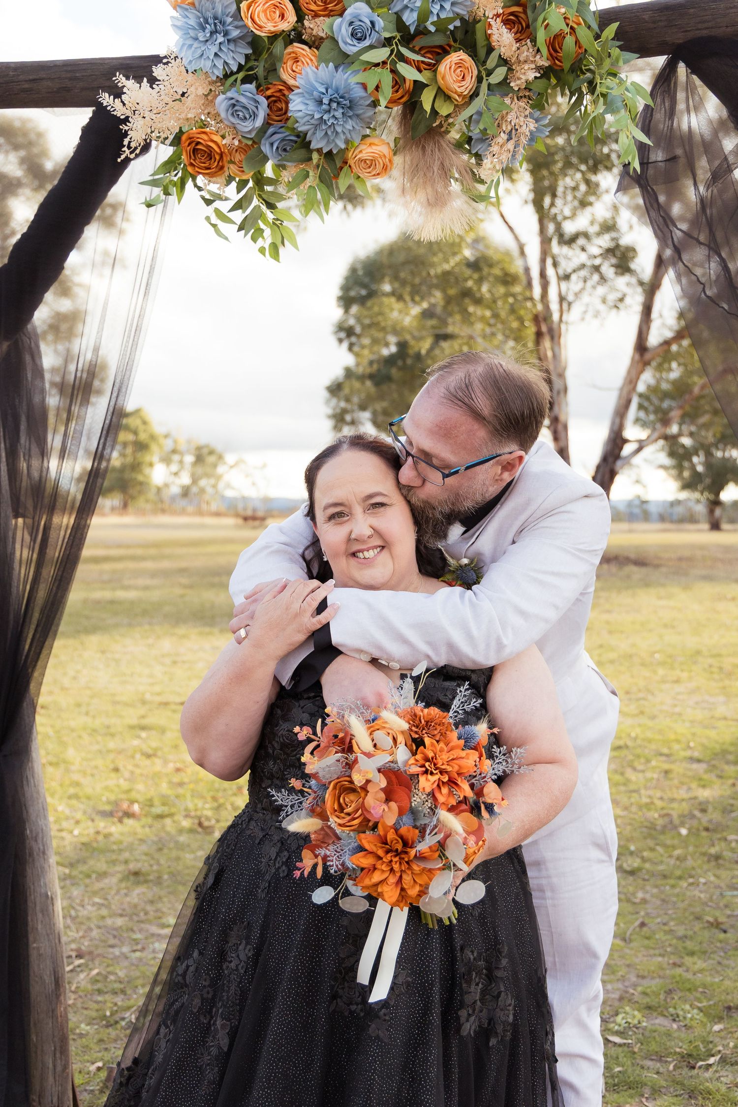 Couple embracing under floral arch at outdoor wedding, bride in black dress holding orange bouquet, groom in white suit kissing her cheek.