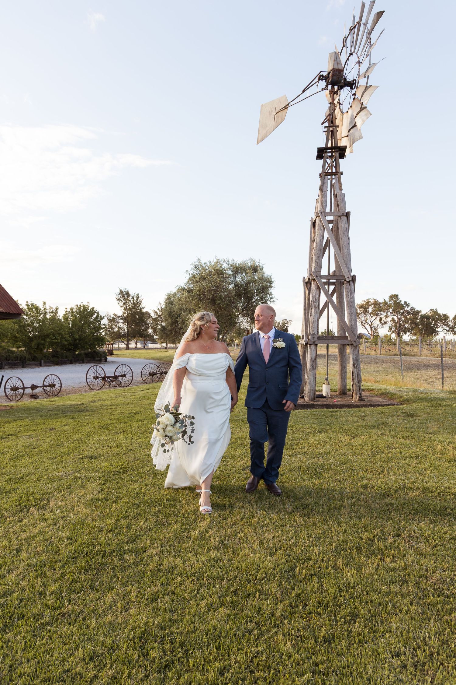 Bride in a white off-the-shoulder gown and groom in a navy suit walk hand in hand on a grassy lawn near a rustic windmill at a wedding.