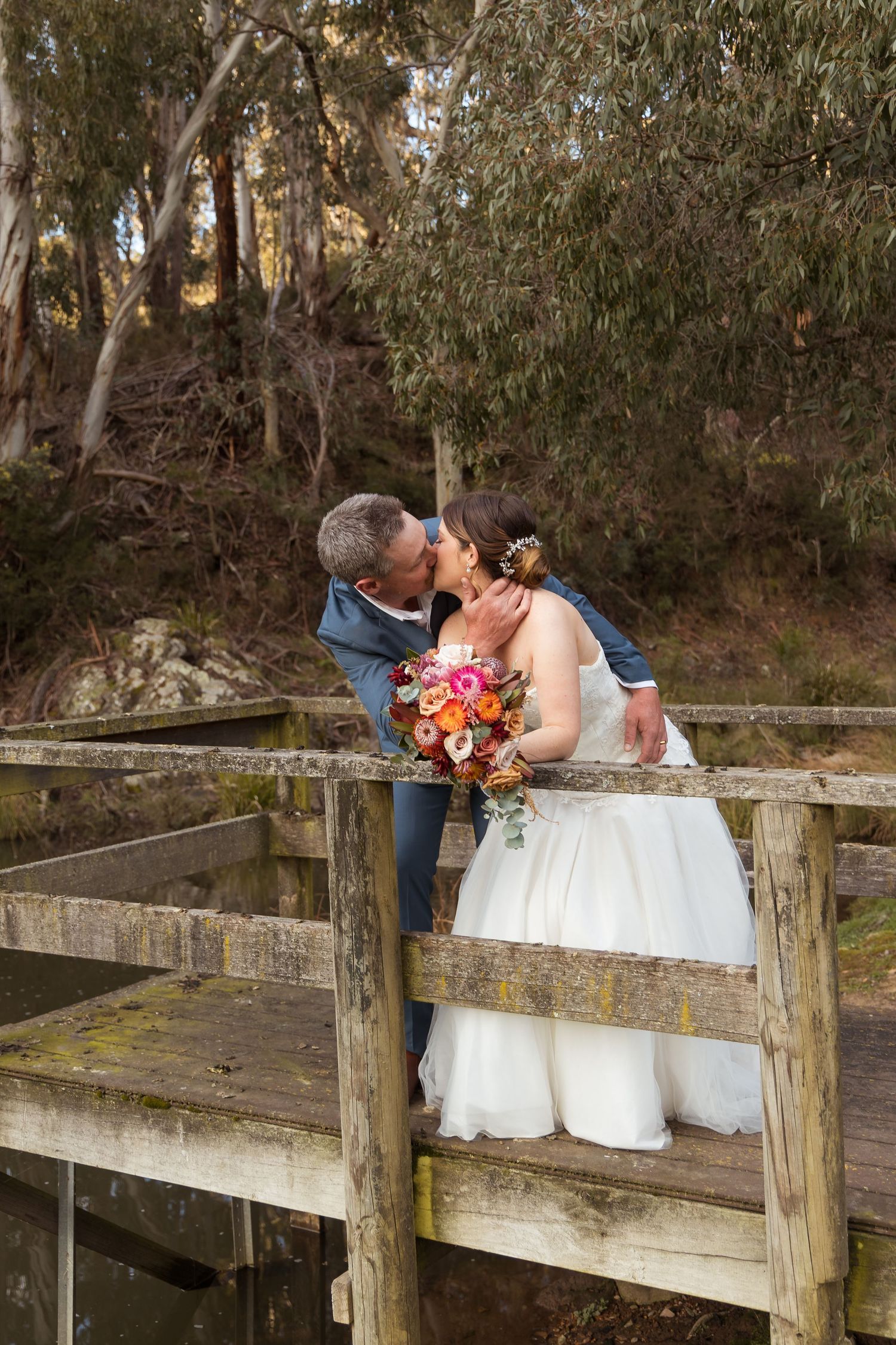 Bride and groom share a romantic kiss on a rustic wooden bridge surrounded by lush trees, bride holding colorful bouquet.