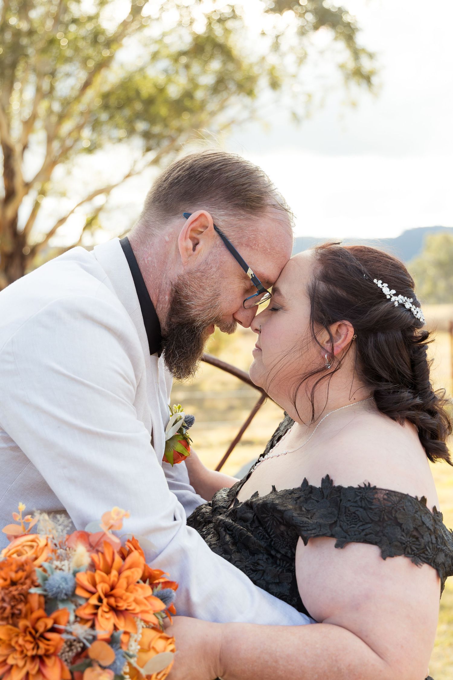 Bride in black lace gown and groom in white suit share romantic forehead touch holding orange floral bouquet outdoors.