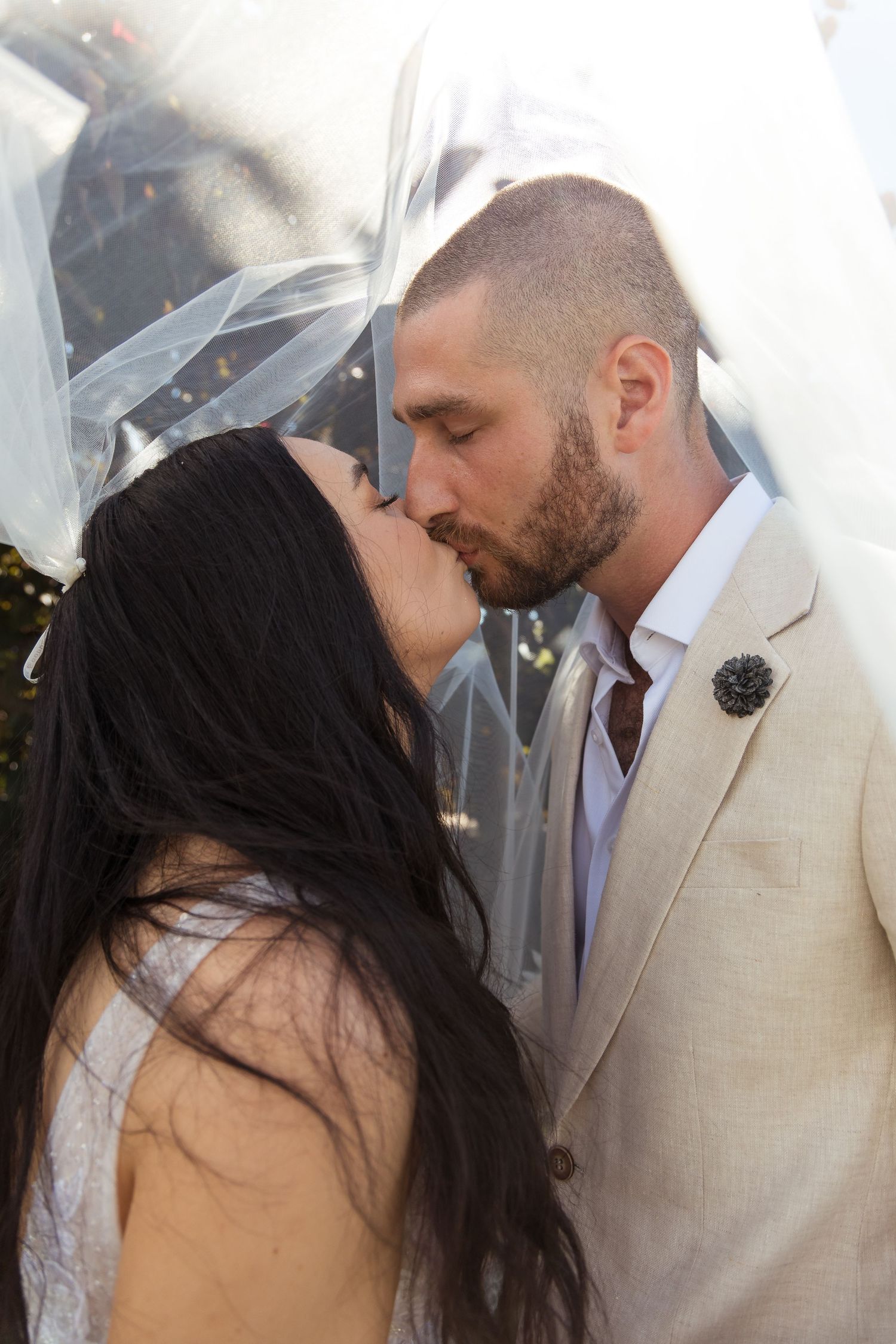Bride and groom sharing romantic kiss under flowing white veil, groom wearing beige suit with boutonniere.