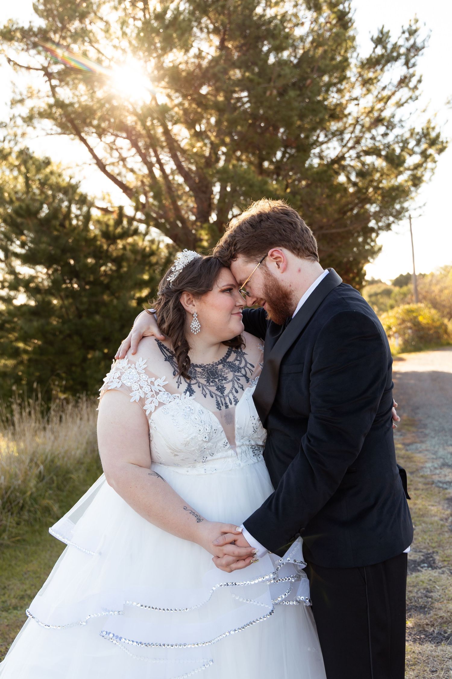 Bride and groom share an intimate forehead touch holding hands, backlit by golden sunset near pine tree.