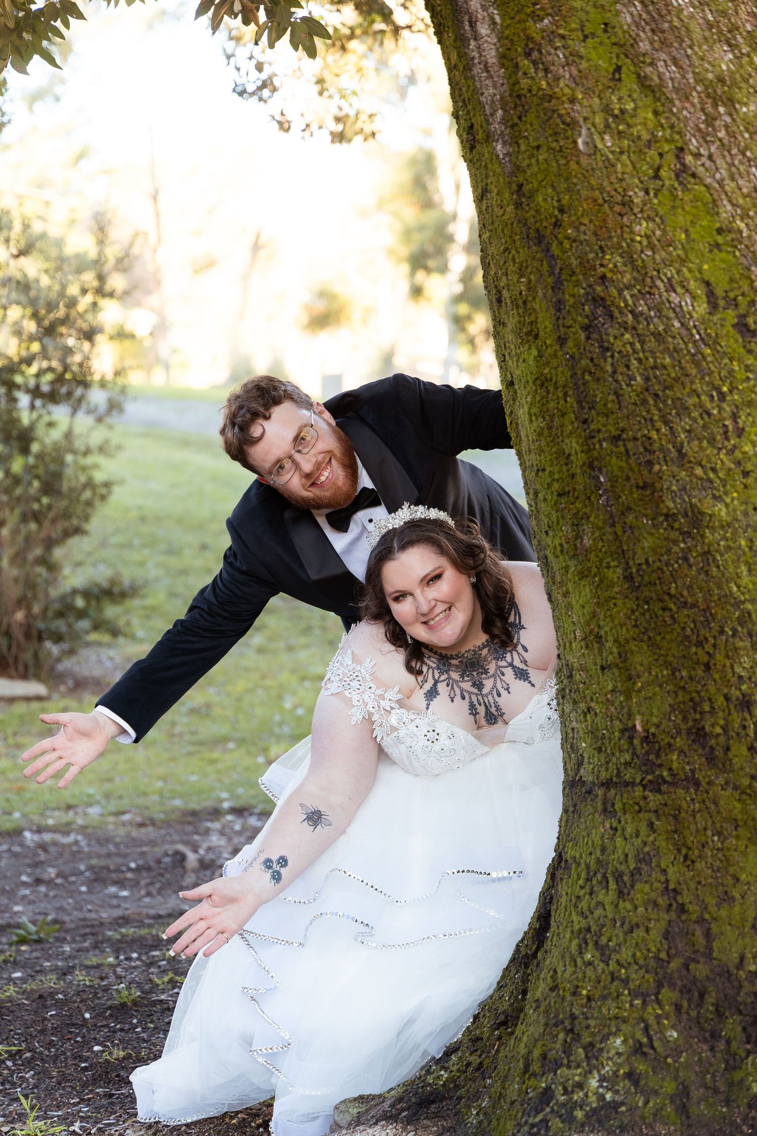 Playful bride and groom pose beside a moss-covered tree, laughing together on their wedding day.