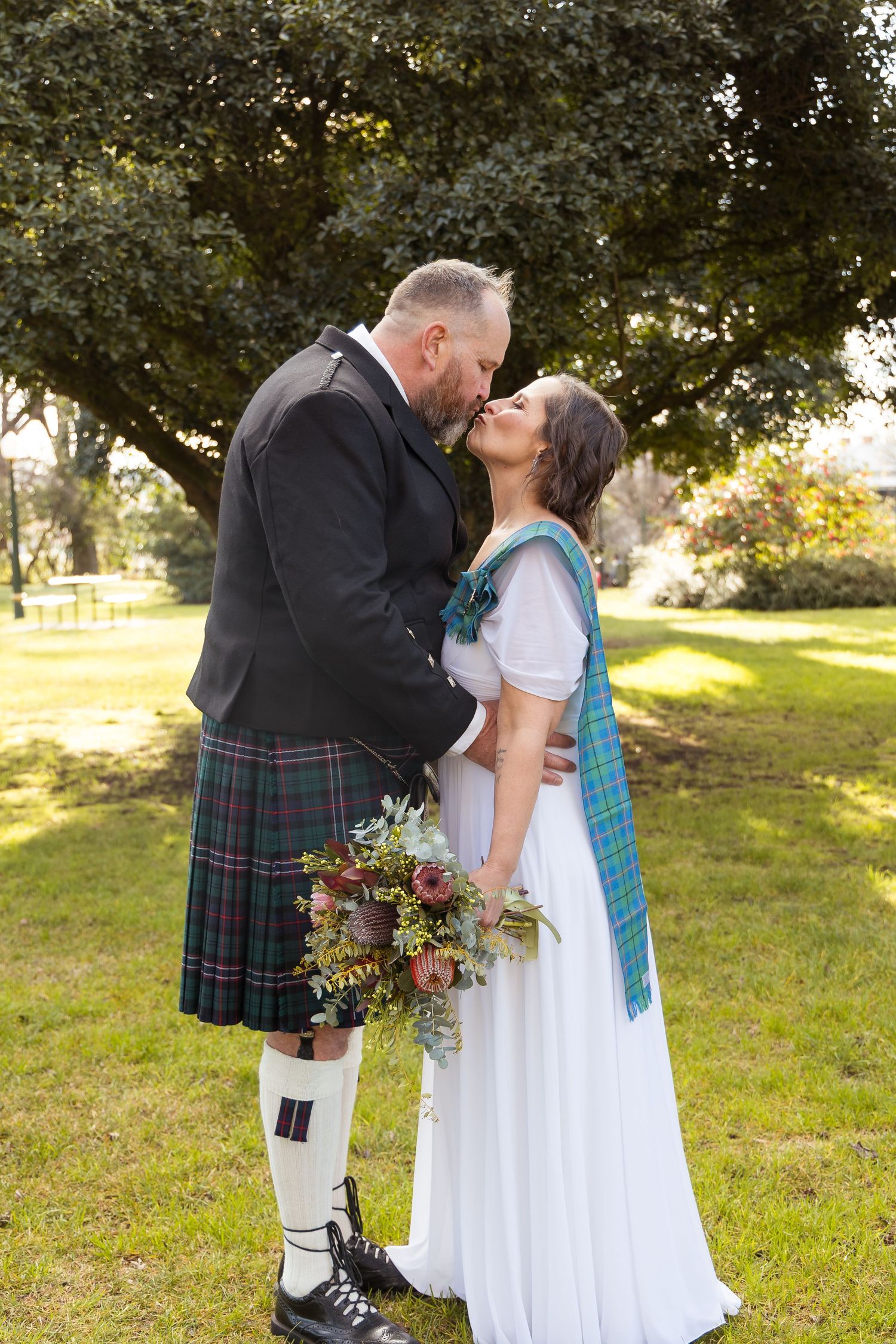 Couple sharing a kiss outdoors in Scottish wedding attire, groom in tartan kilt, bride in white gown with plaid sash.
