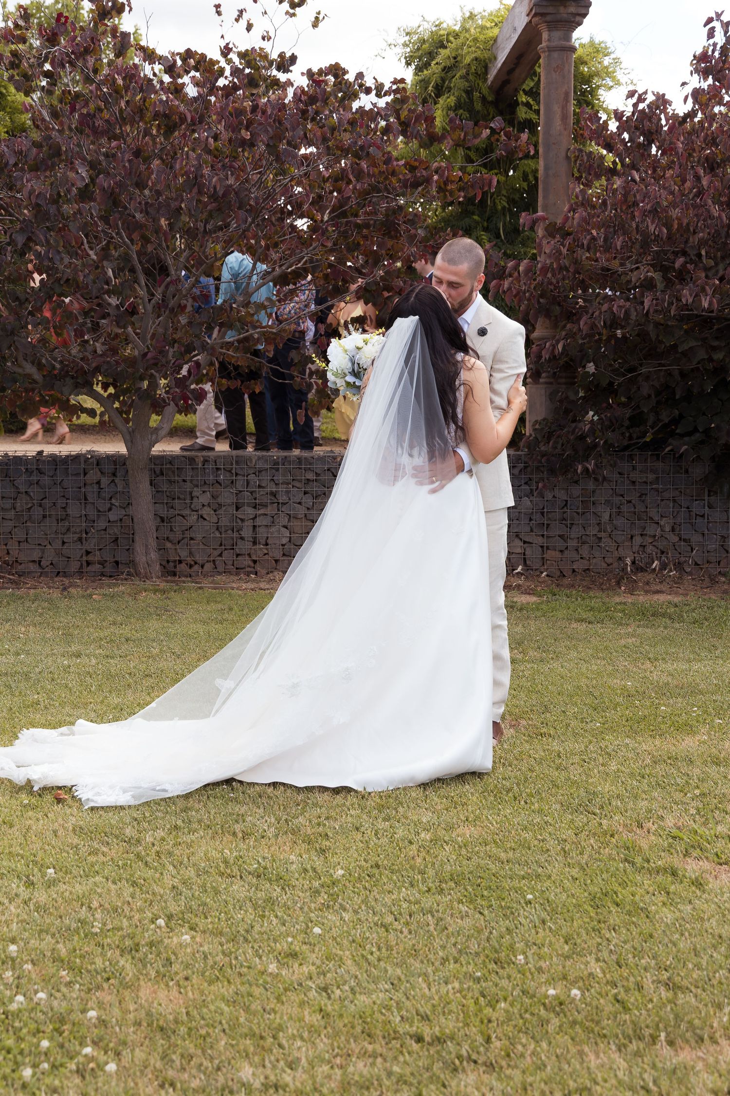 Bride in white gown with cathedral veil embracing groom in cream suit on green lawn with purple foliage.