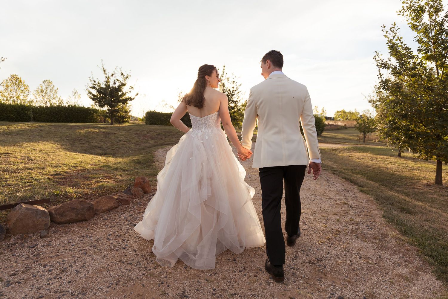 Bride in layered pink gown and groom in white jacket walking hand in hand along scenic outdoor path.