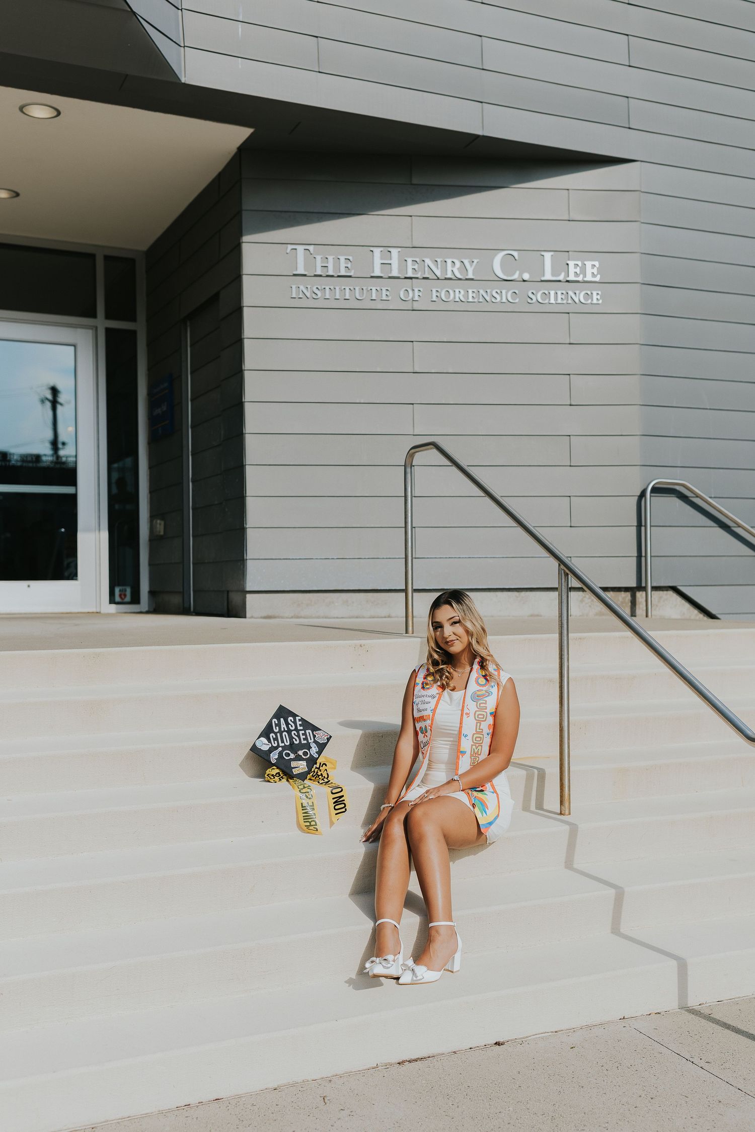 Female graduate in white dress sitting on steps of Henry C. Lee Institute of Forensic Science.