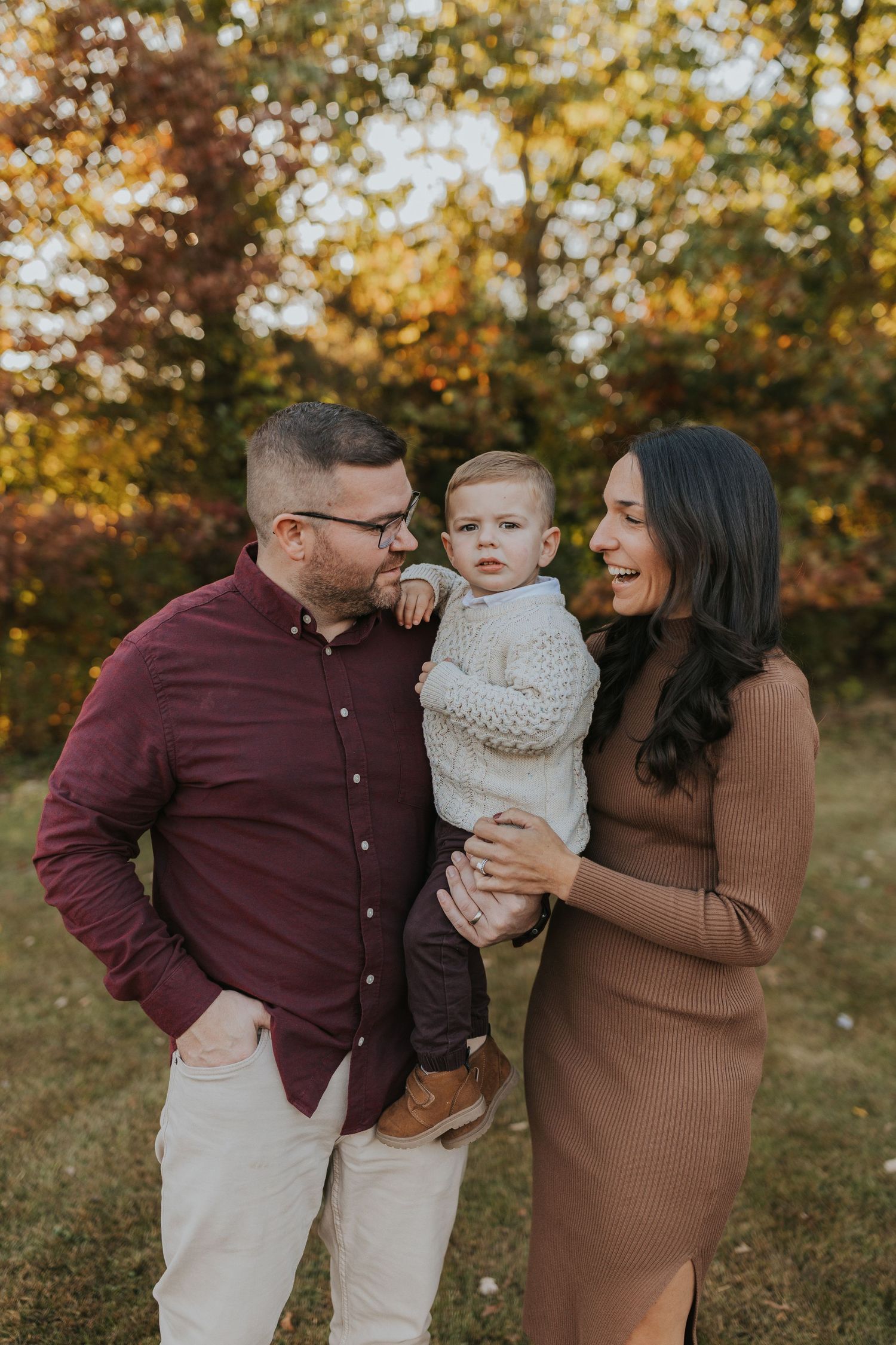 Family of three poses outdoors in fall foliage, parents in burgundy and tan holding their baby boy.