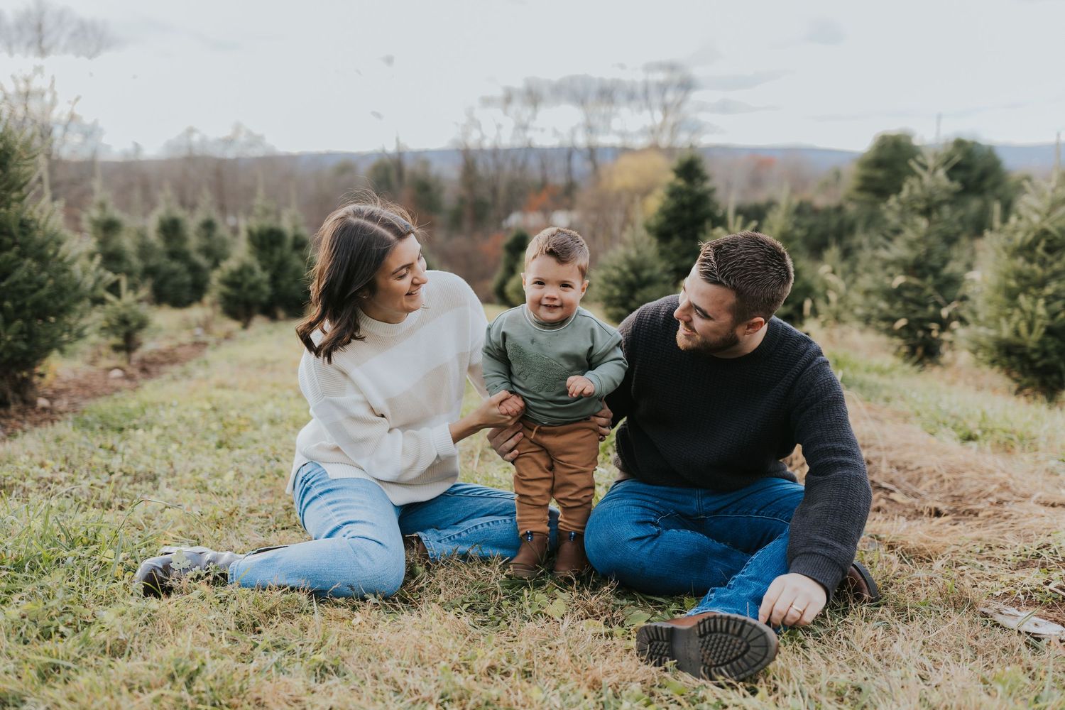 Family of three sitting in a Christmas tree farm field, parents smiling at toddler between them.