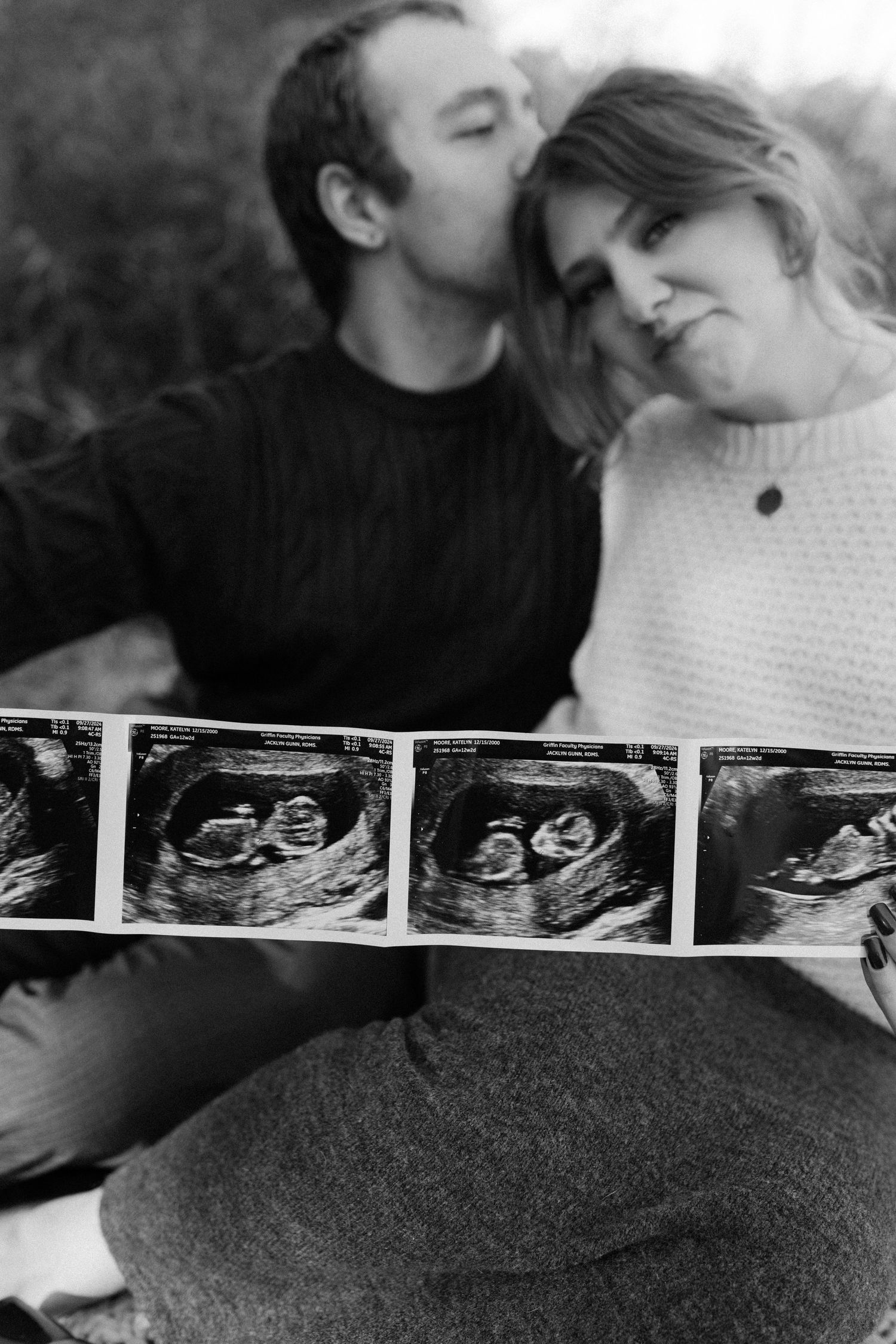 Black and white photo of couple kissing while holding ultrasound images during outdoor maternity session.