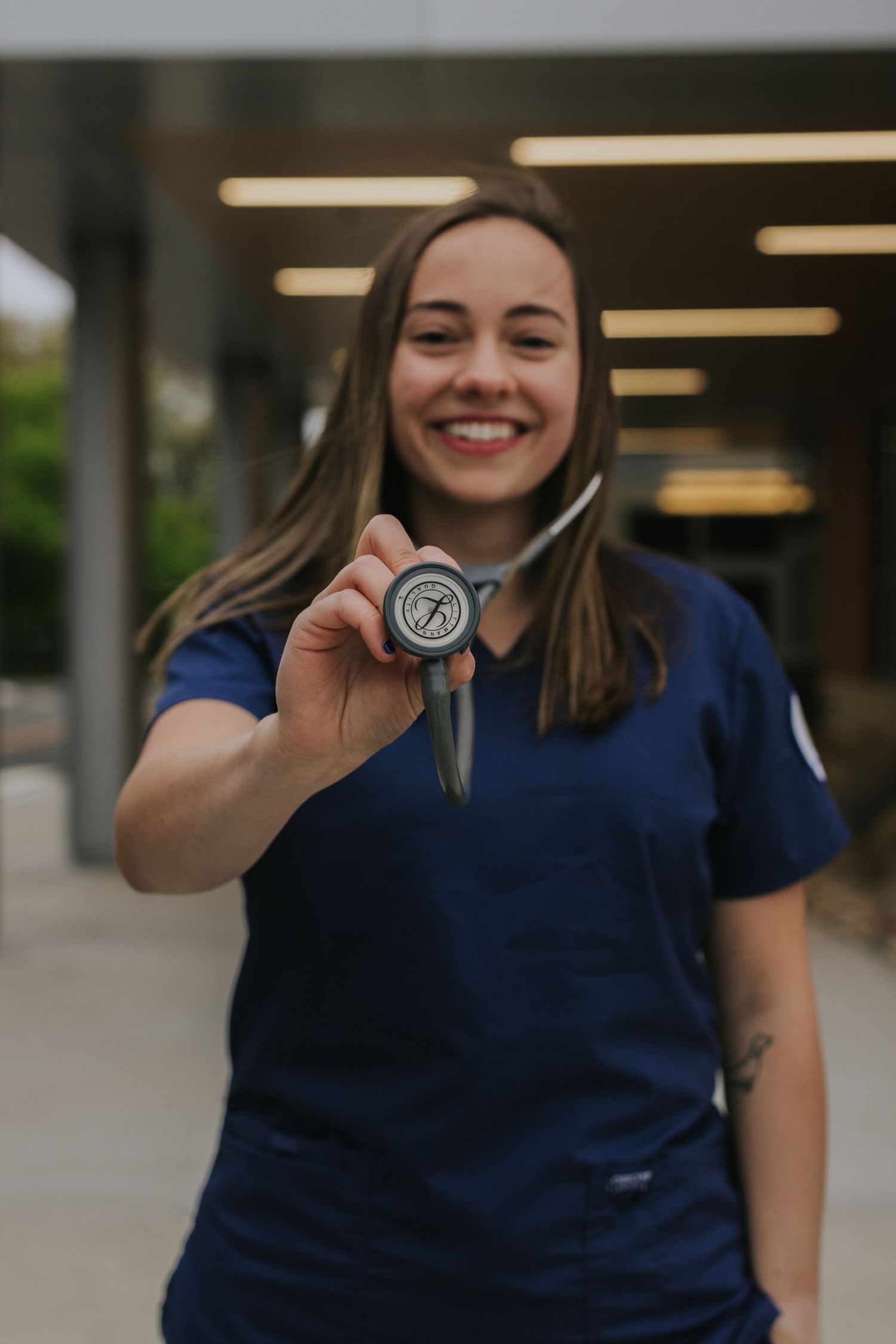 Smiling female nursing student in navy scrubs holds stethoscope toward camera outside university health building.