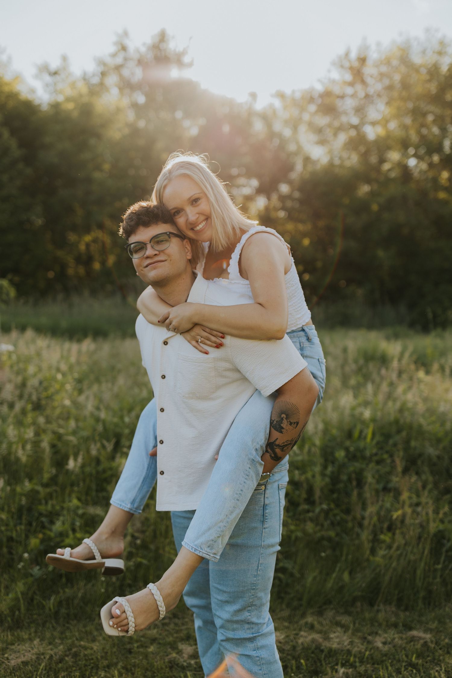 Young man giving smiling blonde woman a piggyback ride in a sunny green meadow at golden hour.
