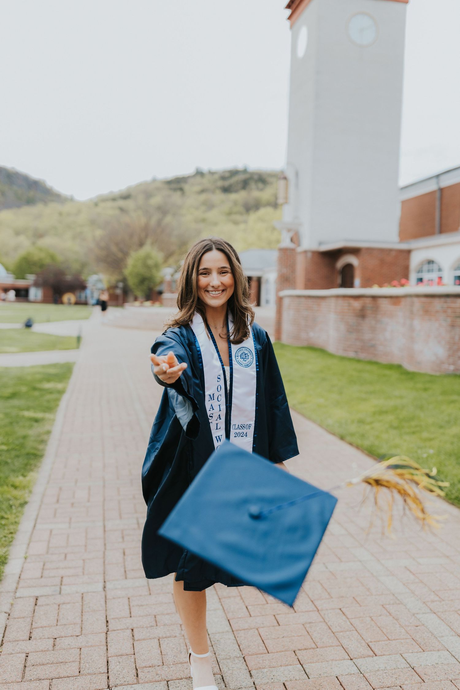 Graduate in navy gown with stole extending cap toward camera on a scenic campus pathway.