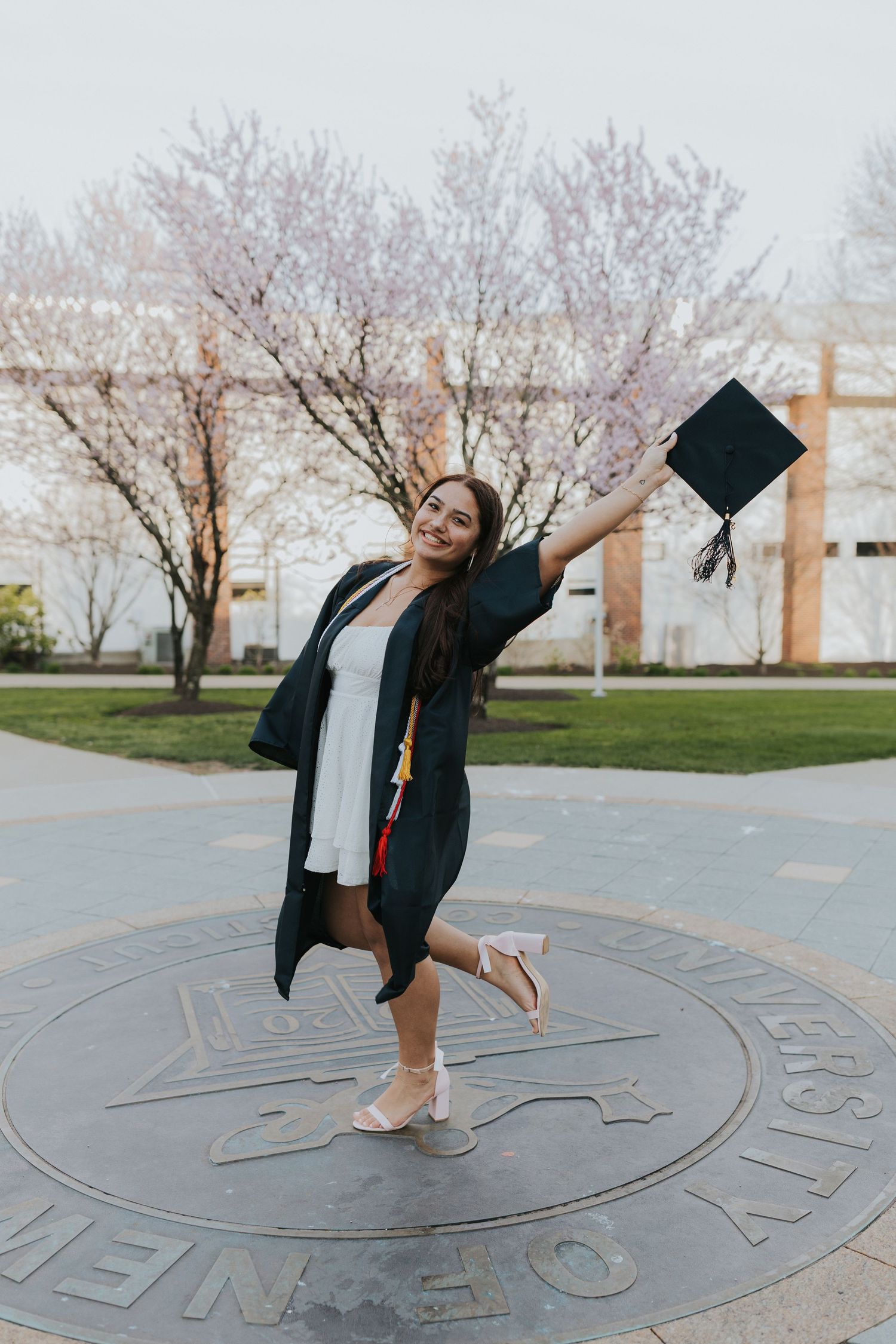 Joyful graduate in cap and gown raising hat while posing on university seal outdoors.