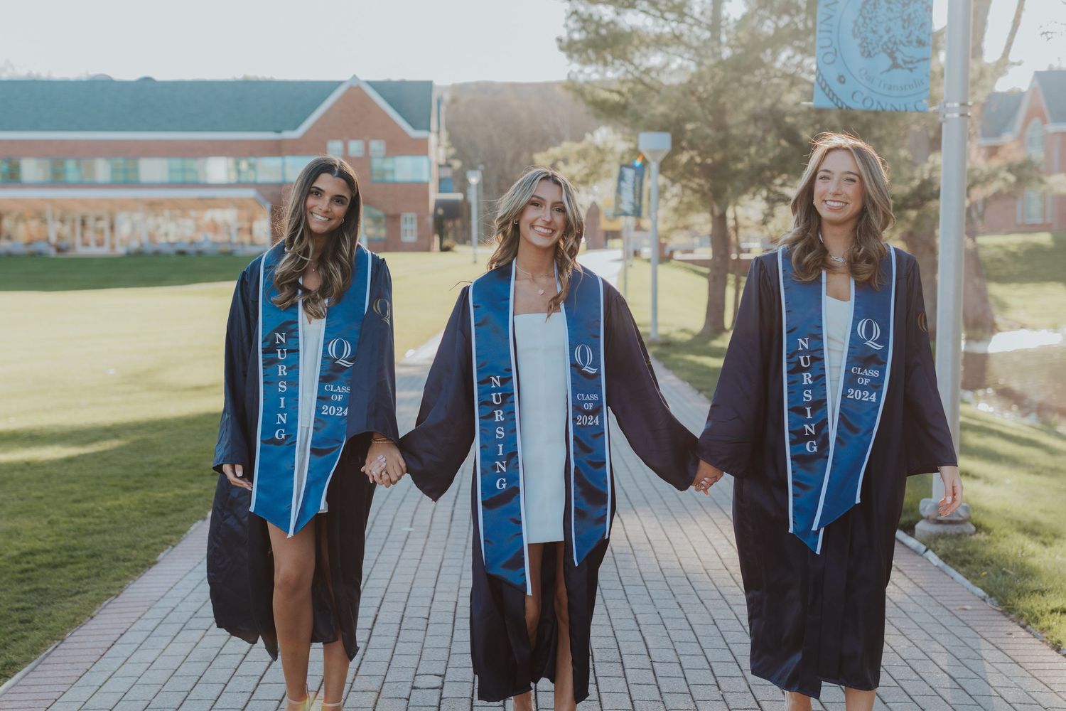 Three graduates in black caps and gowns with blue stoles walking together on campus pathway.