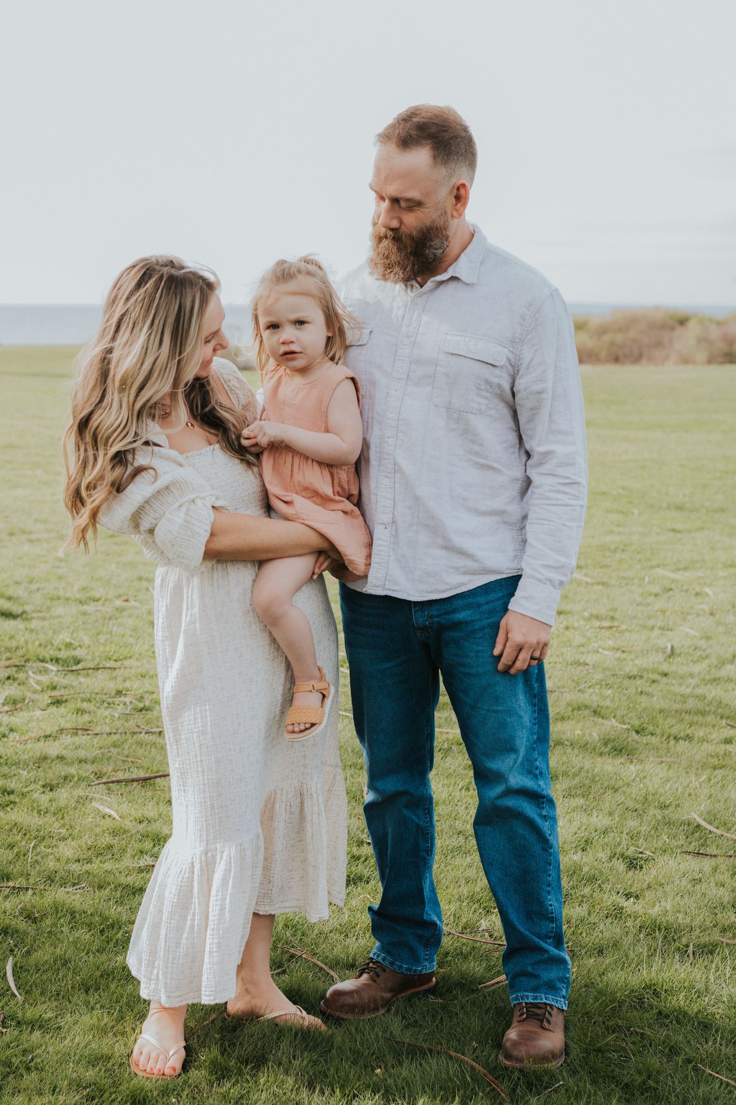 Family of three stands in green meadow, mother in white dress holding toddler girl in pink.