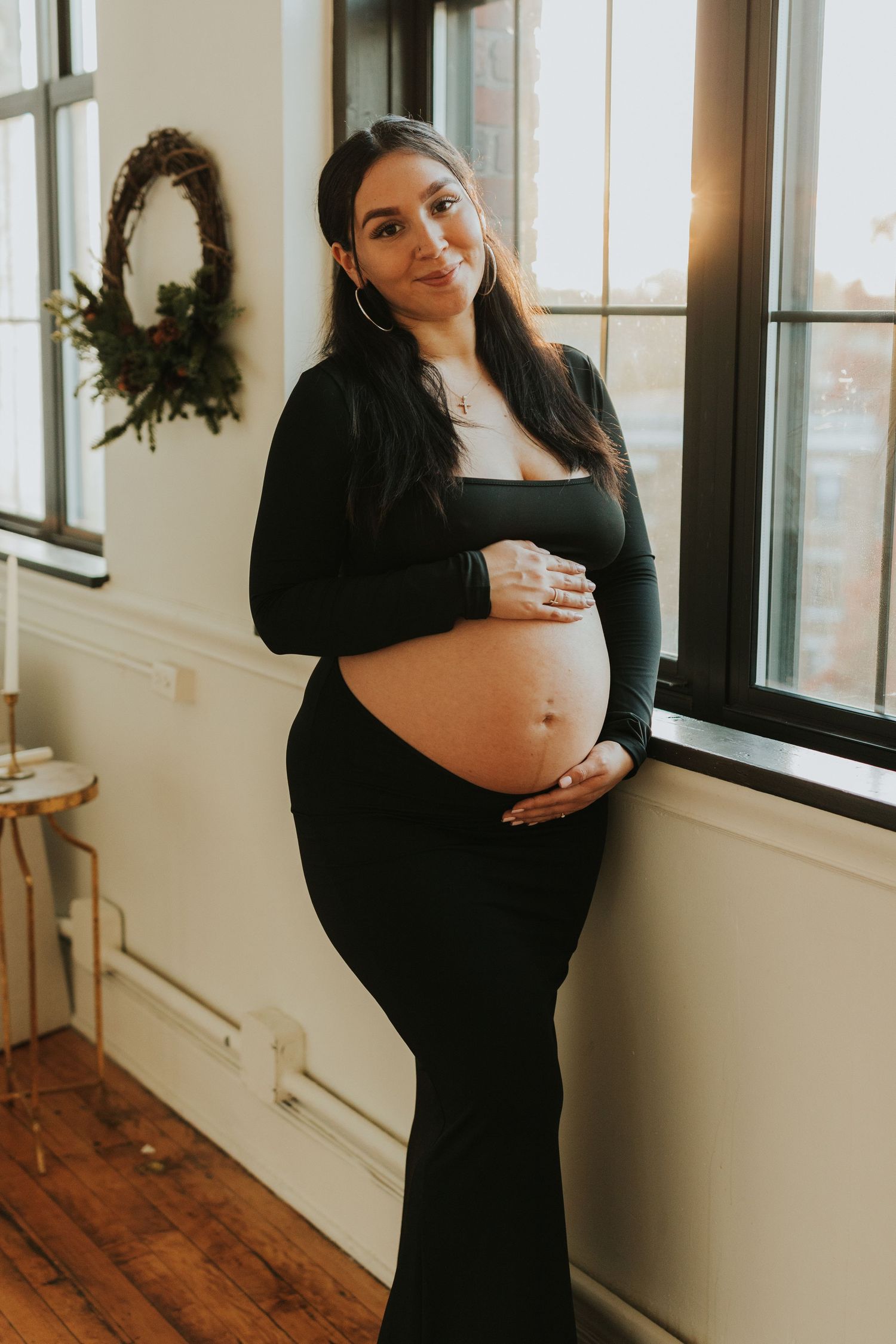 Pregnant woman in black crop top leaning against window, cradling her baby bump.